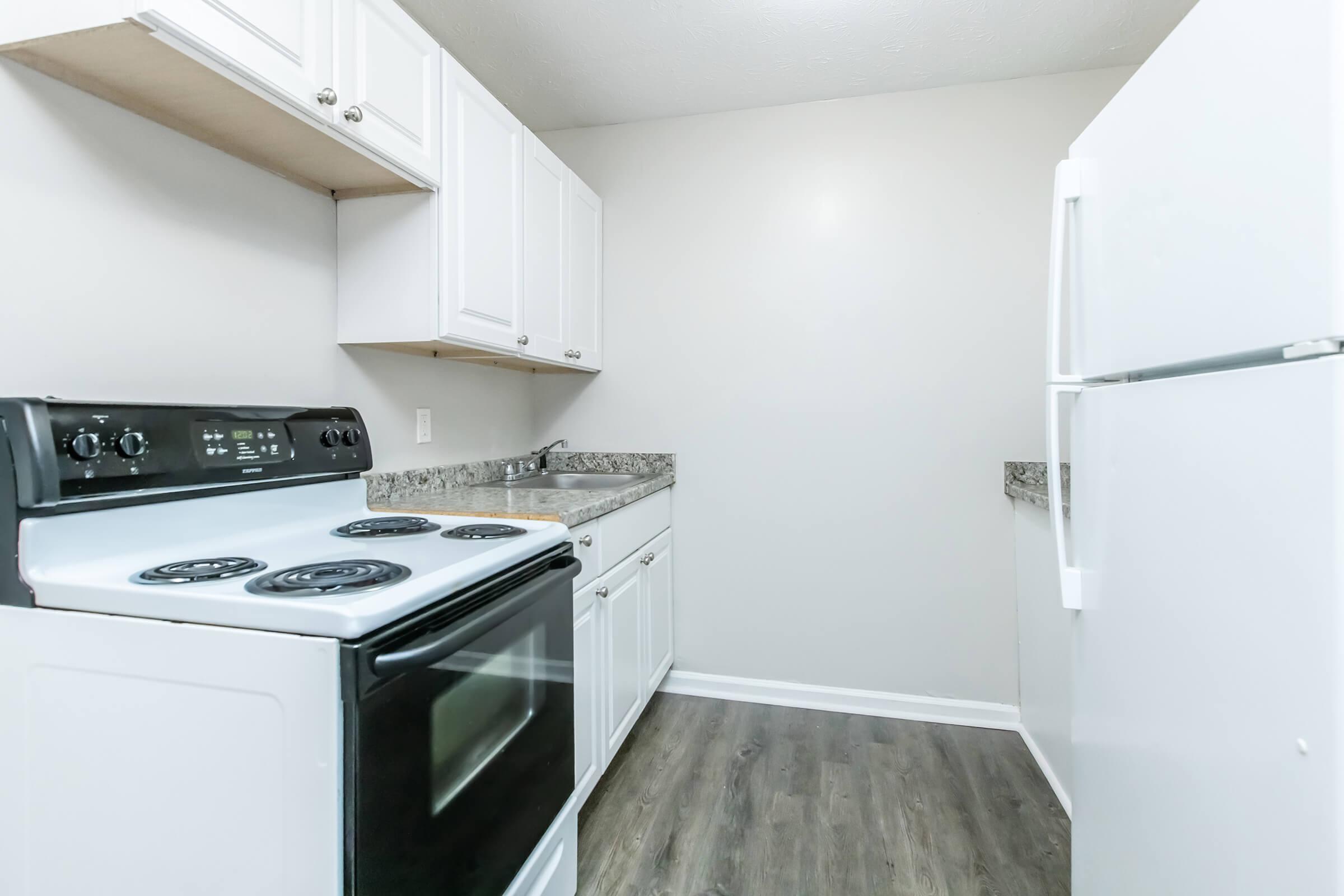 A small kitchen featuring a black stove with four burners, a white refrigerator, and a stainless steel sink. The cabinets are white, and the countertops are a speckled gray. The walls are painted in a light color, and the flooring is dark wood laminate. The kitchen is well-lit and organized.