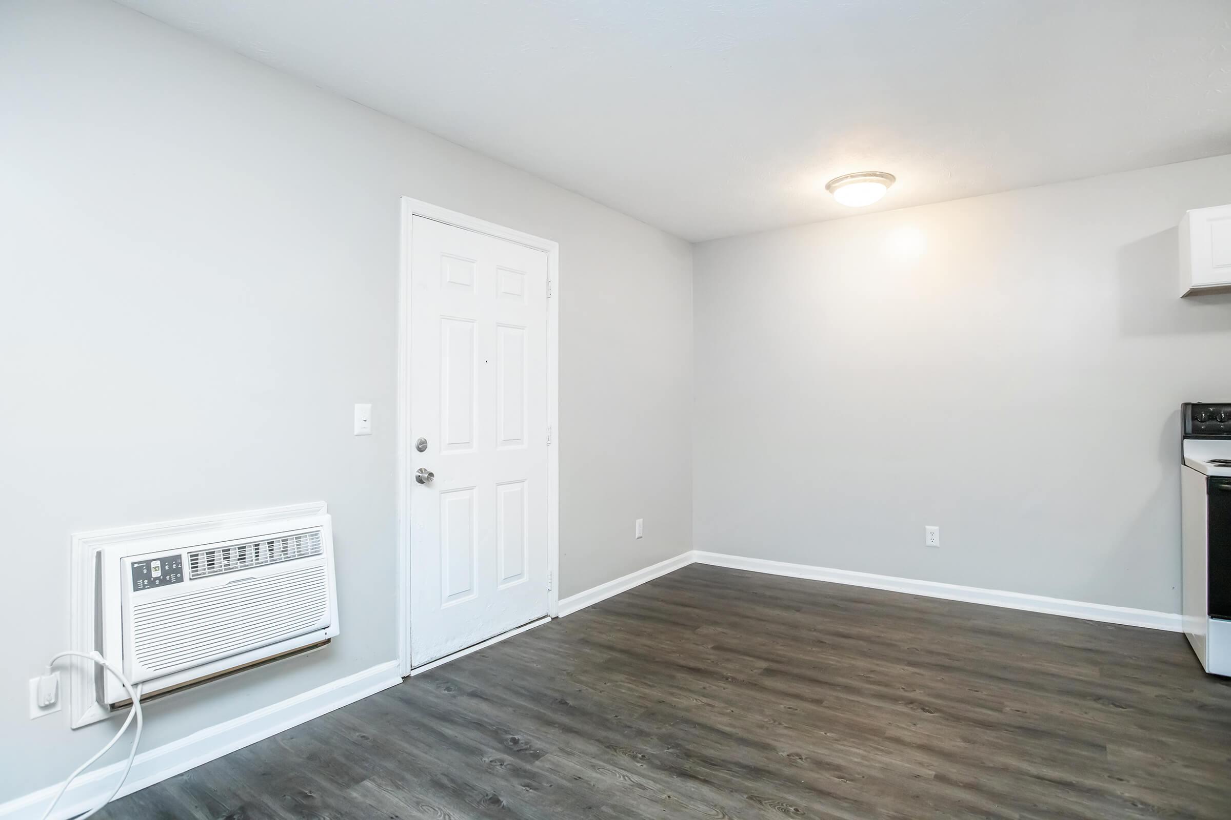 An empty room with light gray walls and a dark wood floor. There's a white door on the left side and an air conditioning unit below it. In the corner, there's a small section of a kitchen with white cabinets. A ceiling light illuminates the space.