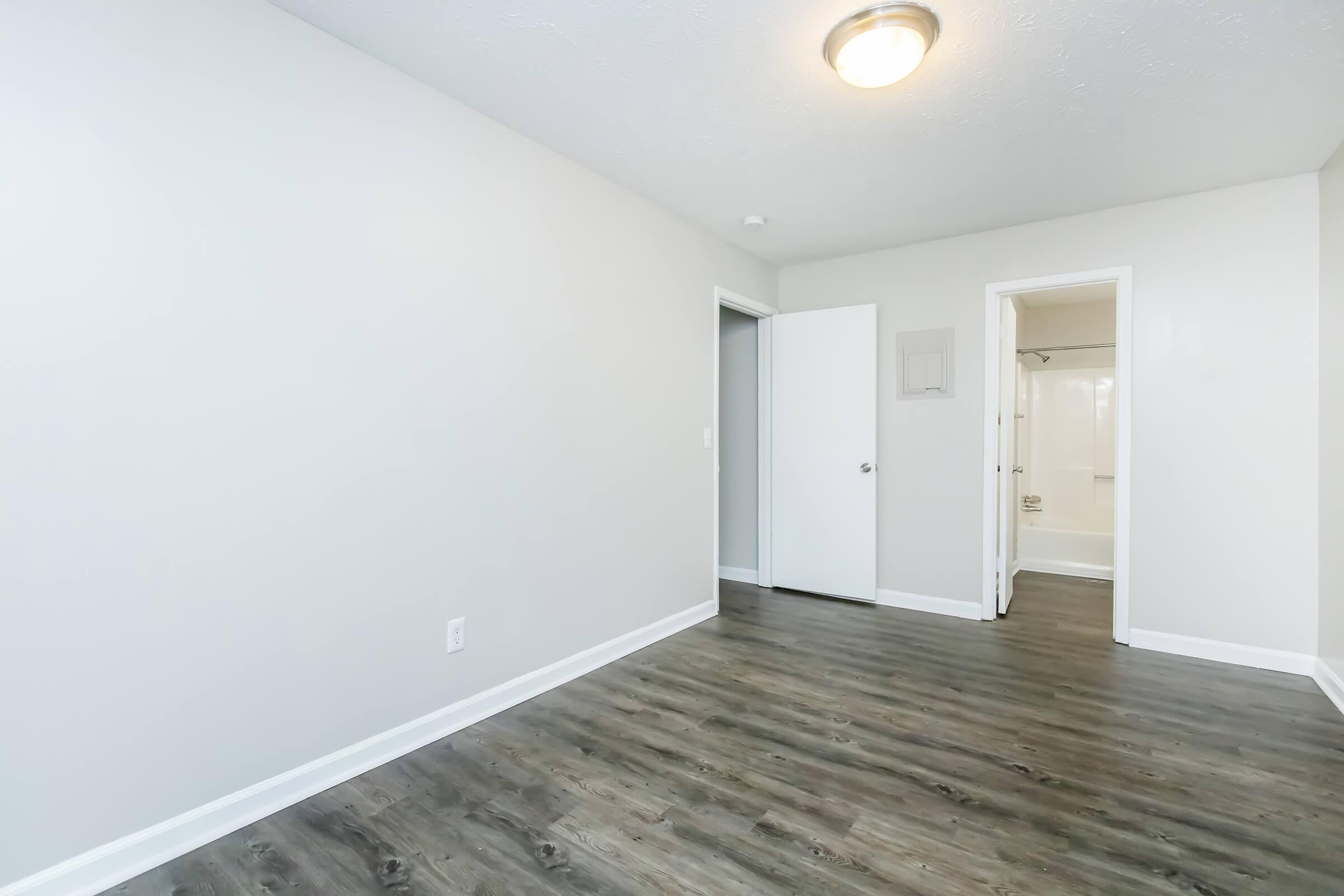 A spacious, empty room with light gray walls and laminate flooring. There is an open doorway leading to a bathroom with a shower, and a closet door on the left. The ceiling has a round light fixture providing illumination.