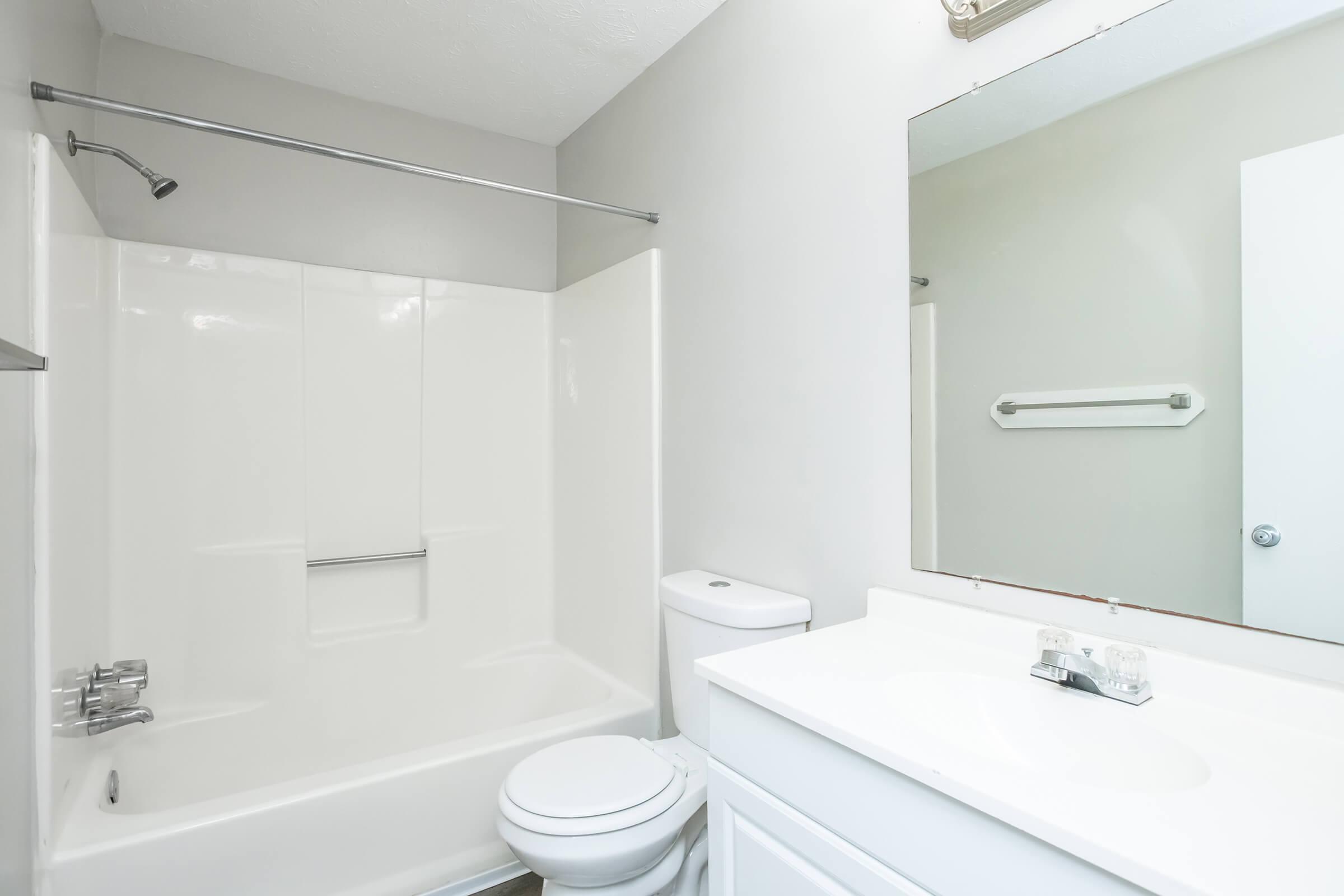 A clean and modern bathroom featuring a white bathtub with shower, a toilet, and a vanity with a sink. The walls are light gray, and there is a large mirror above the sink. The bathroom is well-lit and uncluttered, with a towel bar mounted on the wall.