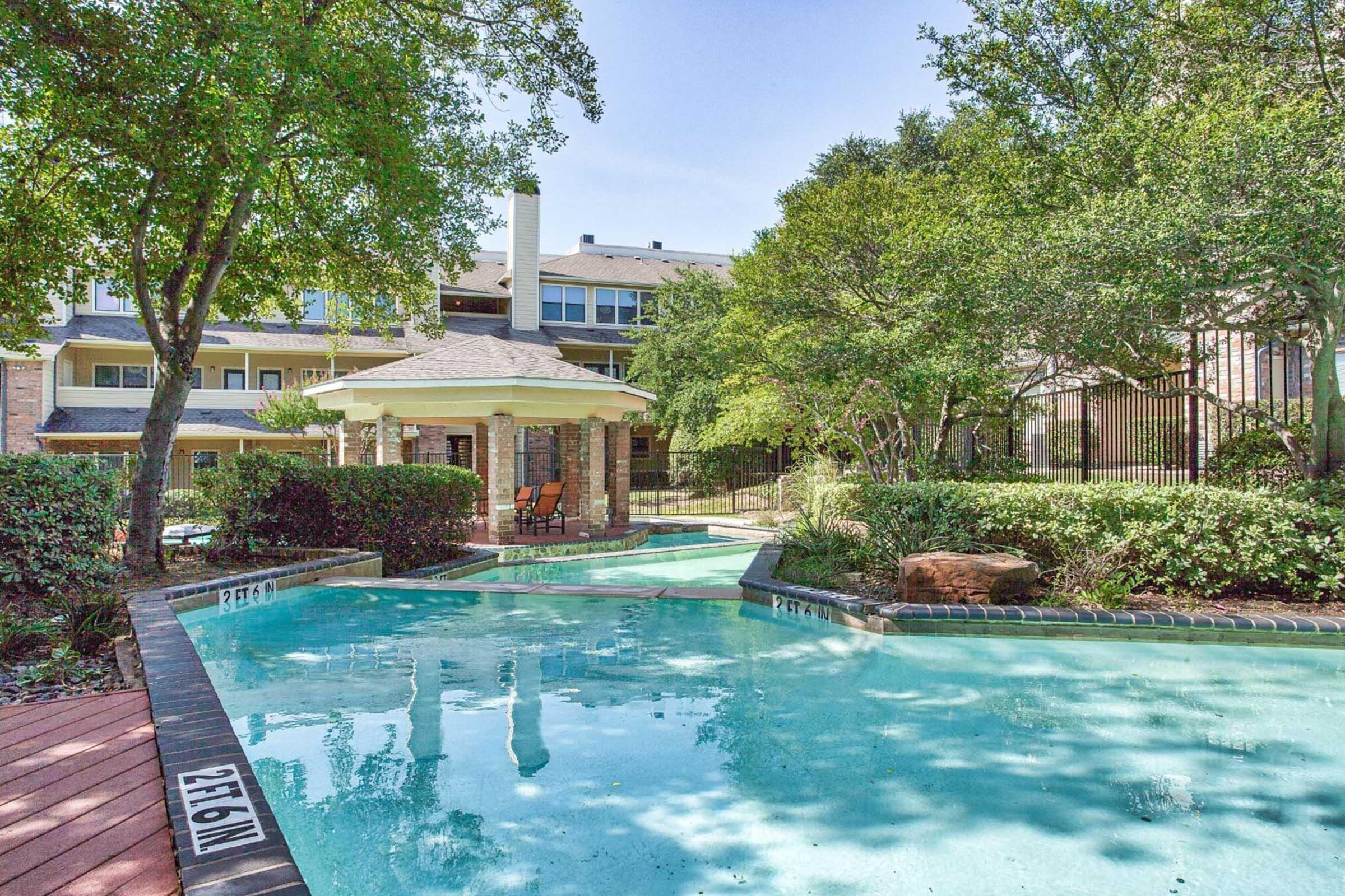 A serene outdoor pool area surrounded by lush greenery, featuring a gazebo with seating. The pool reflects the clear blue sky and is bordered by brick and wooden decking, creating a tranquil atmosphere in a residential complex.