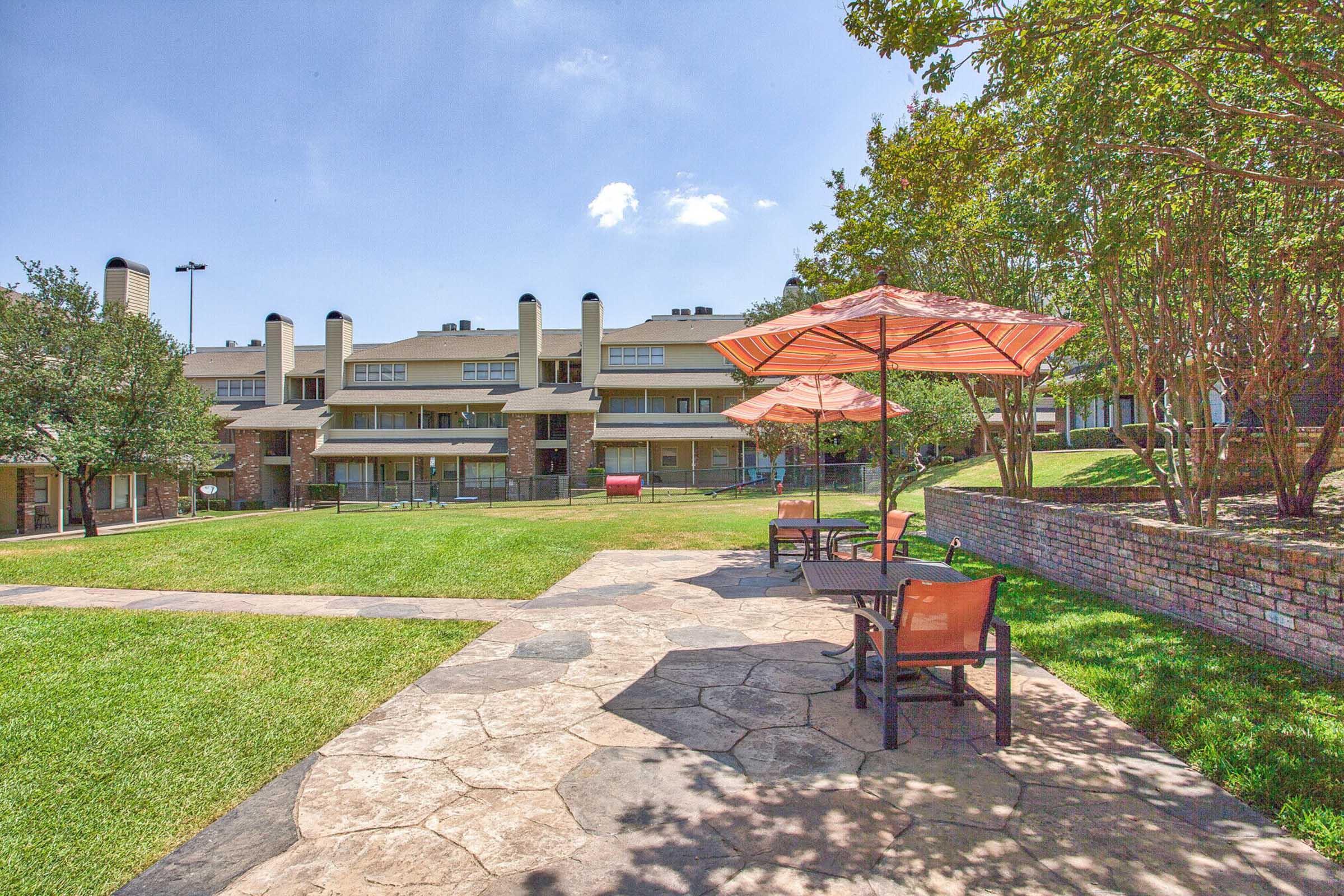 A view of a landscaped area featuring stone pathways, green grass, and patio seating with umbrellas. In the background, there are multi-story buildings with large windows and a clear blue sky above. The scene is bright and inviting, perfect for relaxation outdoors.