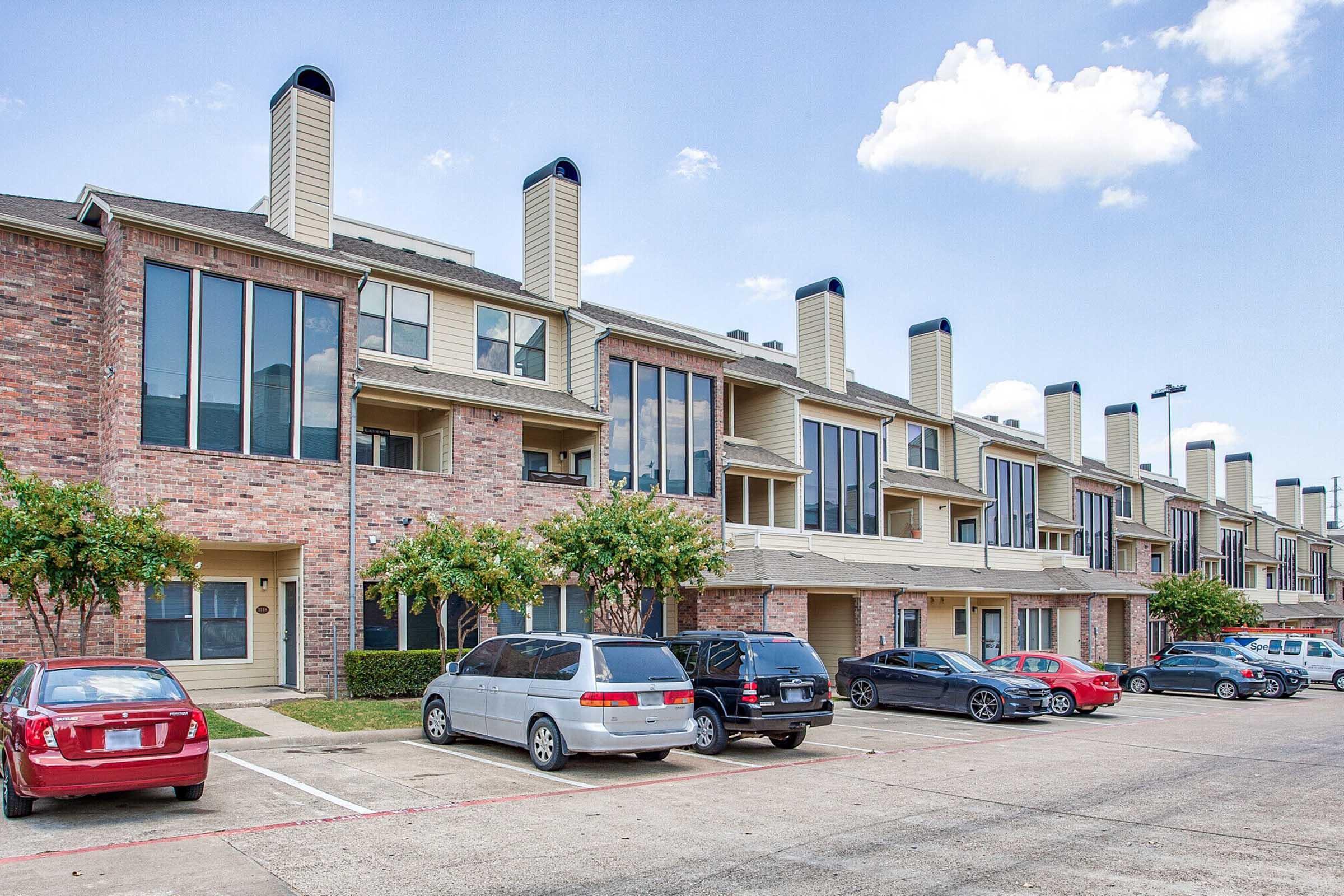 A row of brick apartment buildings with large windows and chimney stacks. Several parked cars are visible in the foreground, and landscaped greenery lines the pathway. The sky is partly cloudy, suggesting a bright day.