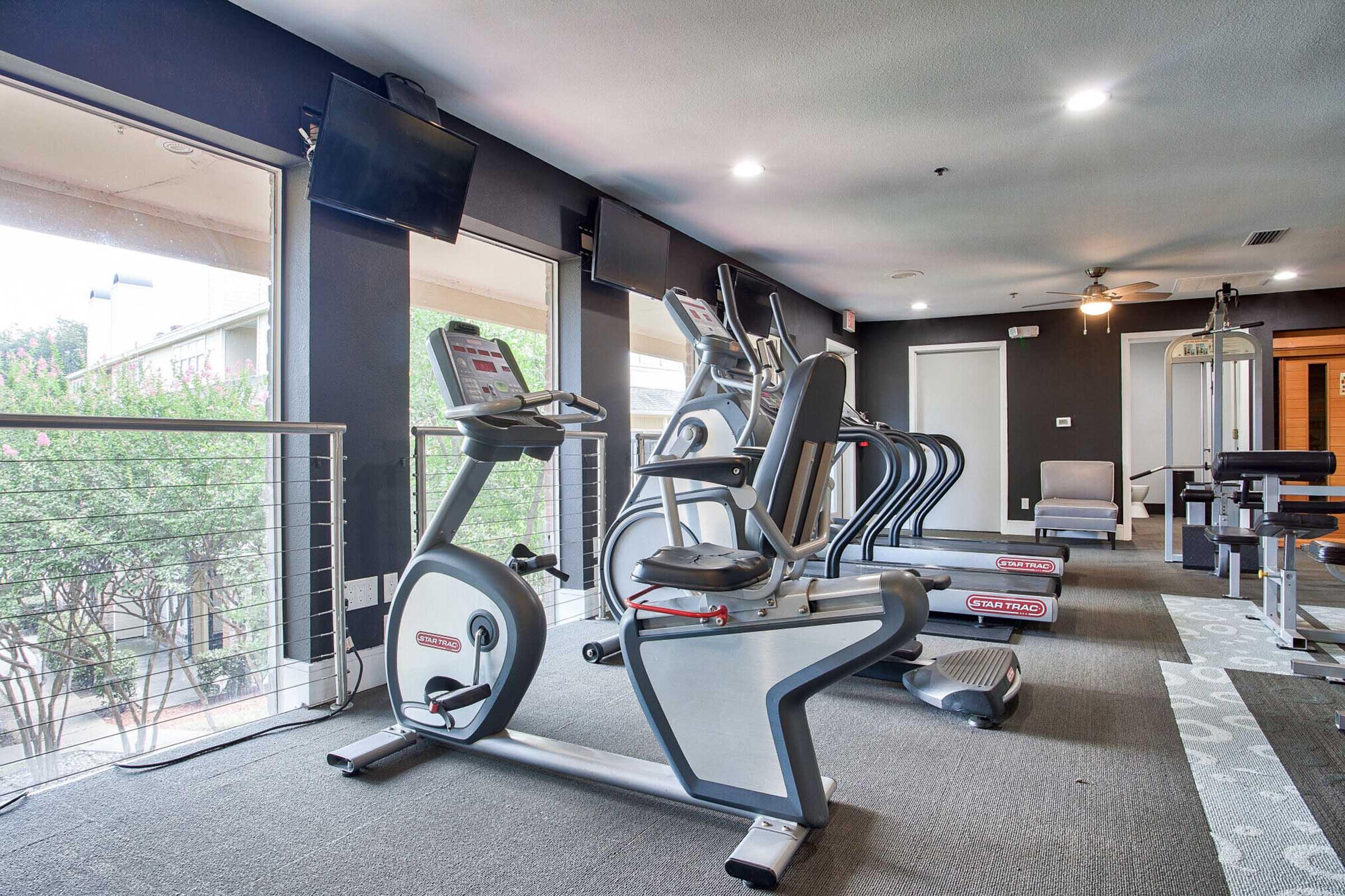A modern gym interior featuring several exercise machines, including stationary bikes and ellipticals. Large windows allow natural light, and multiple flat-screen TVs are mounted on the walls. The floor has a gray carpet, and the space is well-lit with overhead lighting.