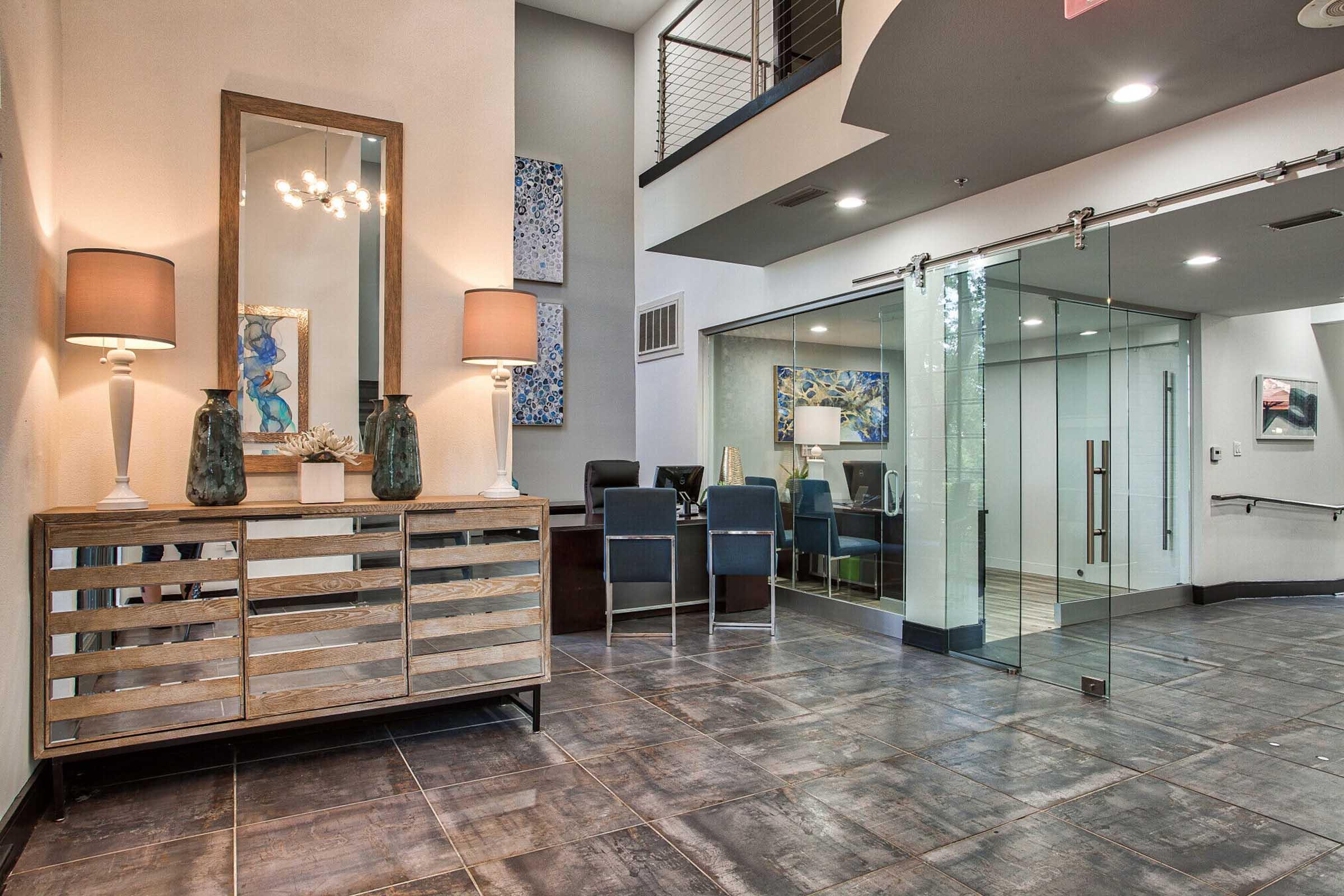Modern lobby featuring a mirrored console table with decorative lamps and vases. Abstract art is displayed on the walls, and there are glass doors leading to a meeting area. The flooring is dark tile, and the space is well-lit, creating a welcoming atmosphere.