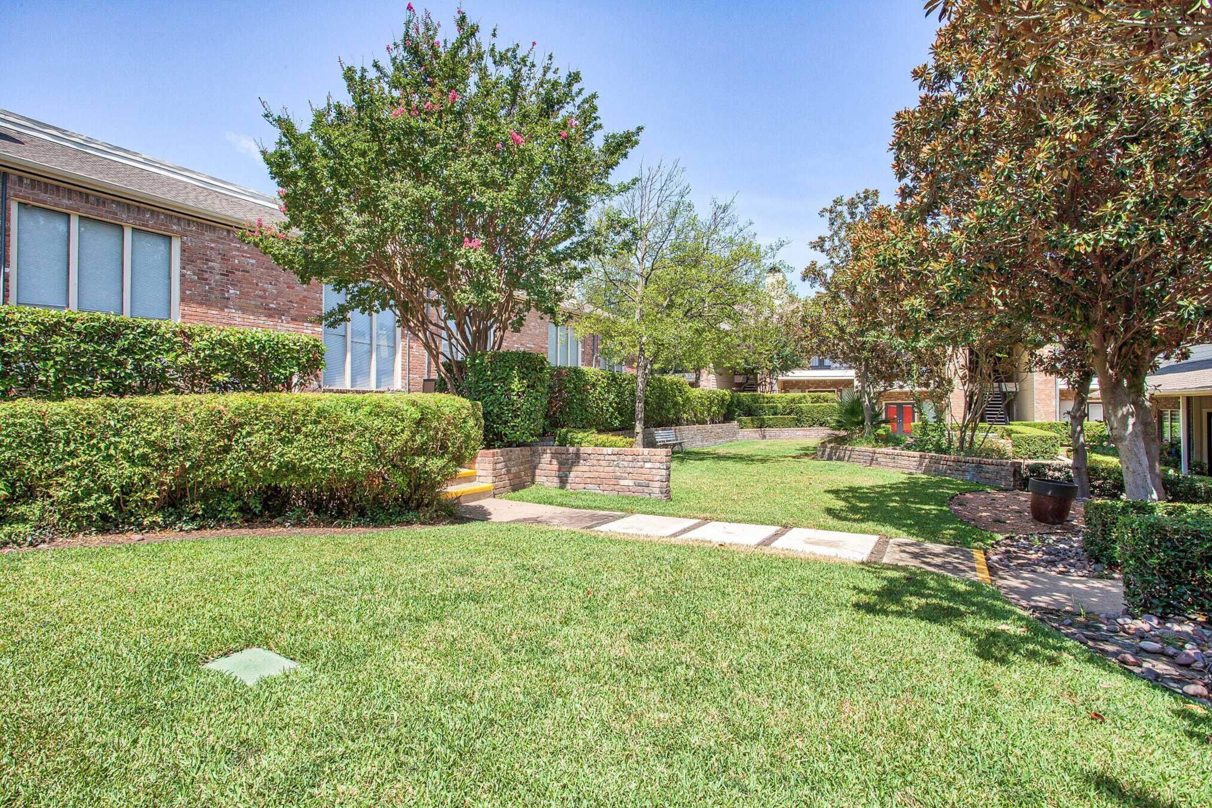 A well-maintained residential area featuring green grass, flowering trees, and neatly trimmed hedges. Pathways lead through the landscape to brick buildings. The setting is bright and sunny, suggesting a warm day.