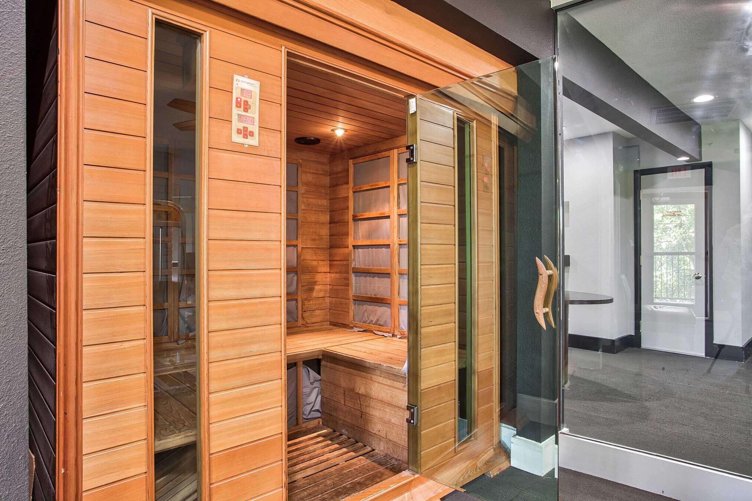 An indoor sauna made of wood with double glass doors, offering a warm and inviting space for relaxation. The interior features wooden benches and a soft light, creating a tranquil atmosphere. The sauna is part of a modern wellness area, visible through a sleek glass partition.