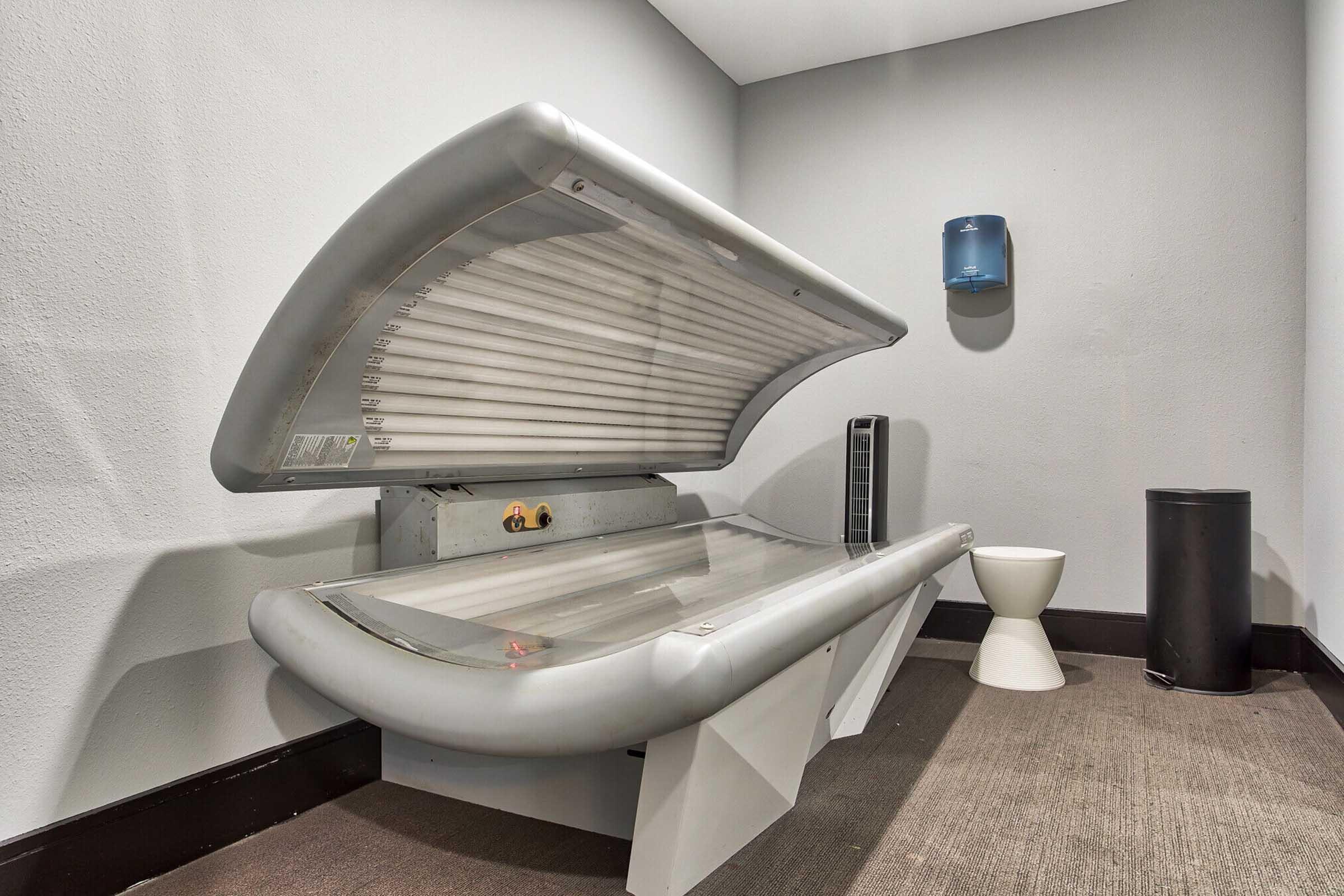 A tanning bed with a curved top, positioned against a plain gray wall, accompanied by a small stool, a trash can, and a wall-mounted paper towel dispenser. The floor is covered with carpet, creating a simple, functional space for indoor tanning.