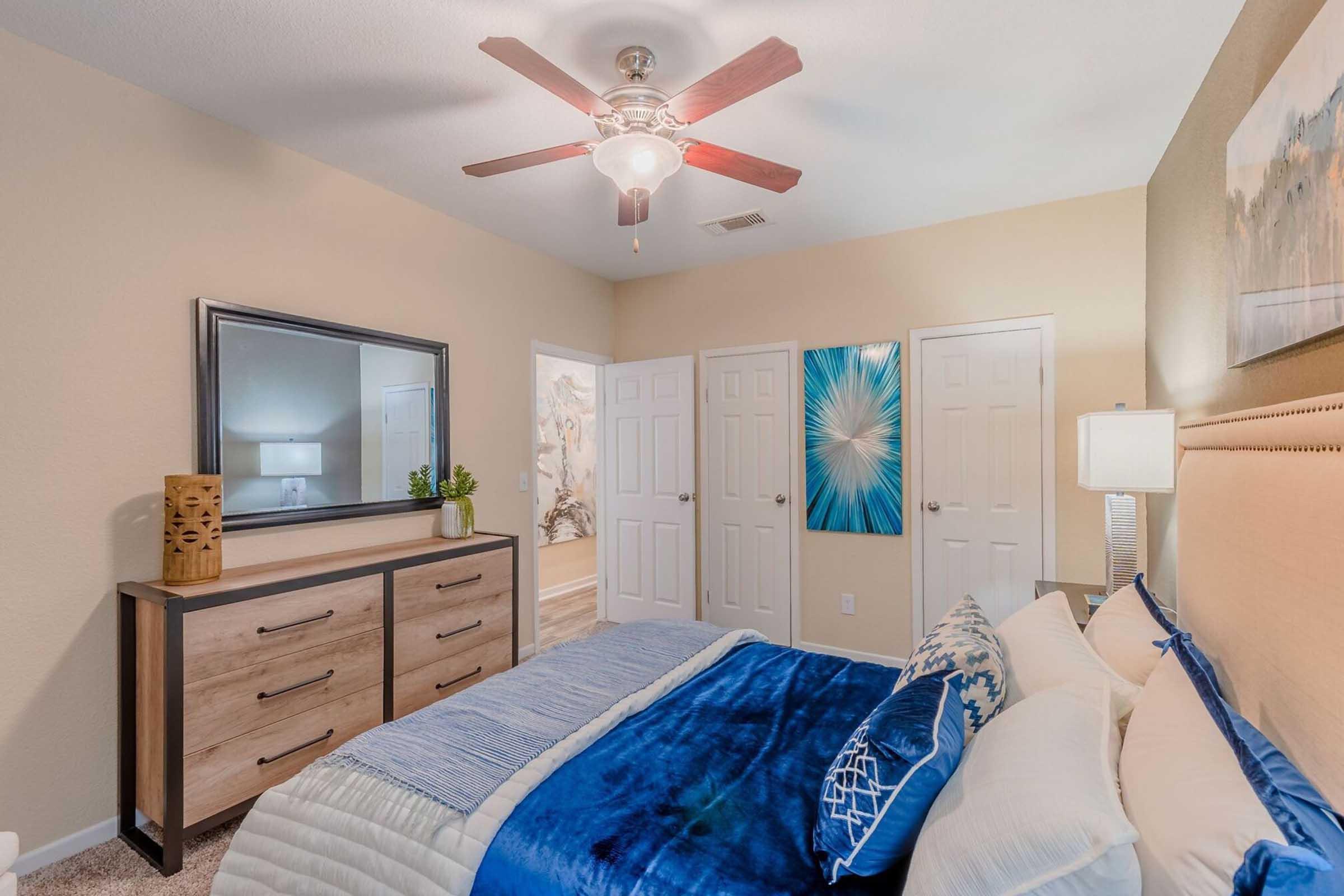 A cozy bedroom featuring a large bed with a blue and white duvet, a wooden dresser, a decorative lamp, and a ceiling fan. The walls are painted in a warm neutral tone, and there are two framed blue artworks on the wall, along with white double doors leading to another room.
