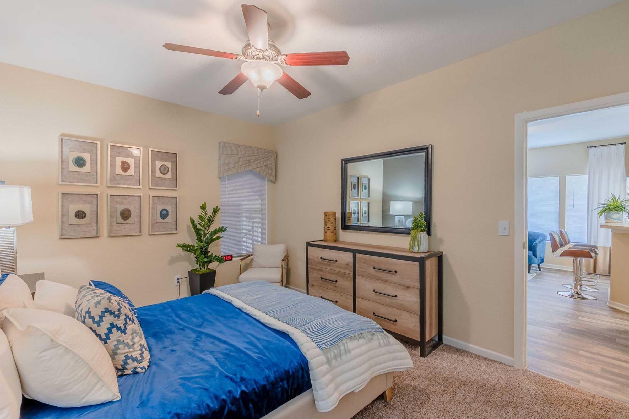 A cozy bedroom featuring a blue bedspread with decorative pillows, a wooden dresser, framed artwork on the wall, and a ceiling fan. Natural light enters through the window, and a doorway leads to another room with a seating area. Green plants add a touch of nature to the space.