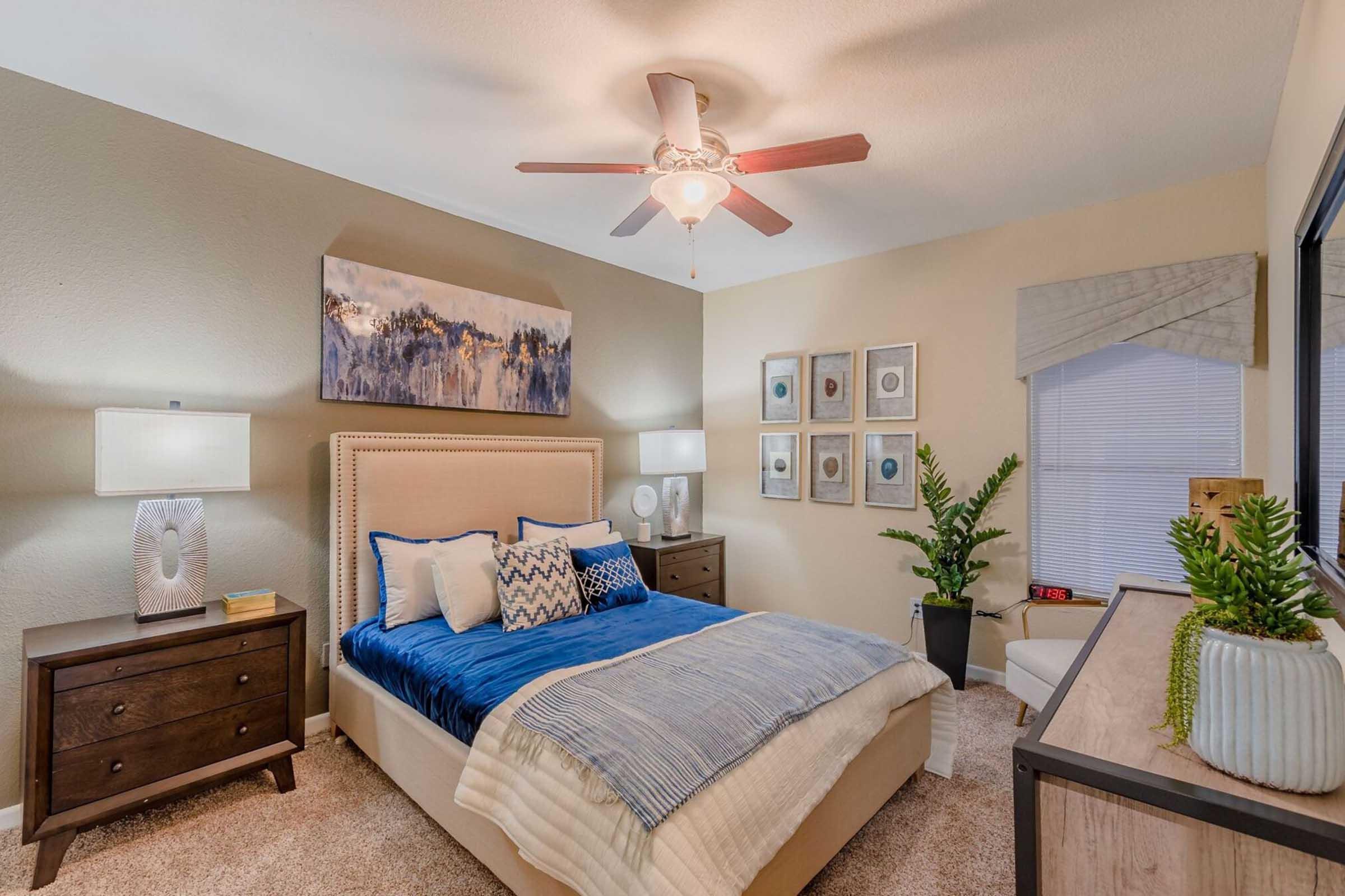 A cozy bedroom featuring a large bed with blue and white bedding, bedside lamps on wooden nightstands, and a ceiling fan. There are decorative wall art and a potted plant, with natural light coming through a window covered by a light shade. The room has neutral-colored walls and carpeting.