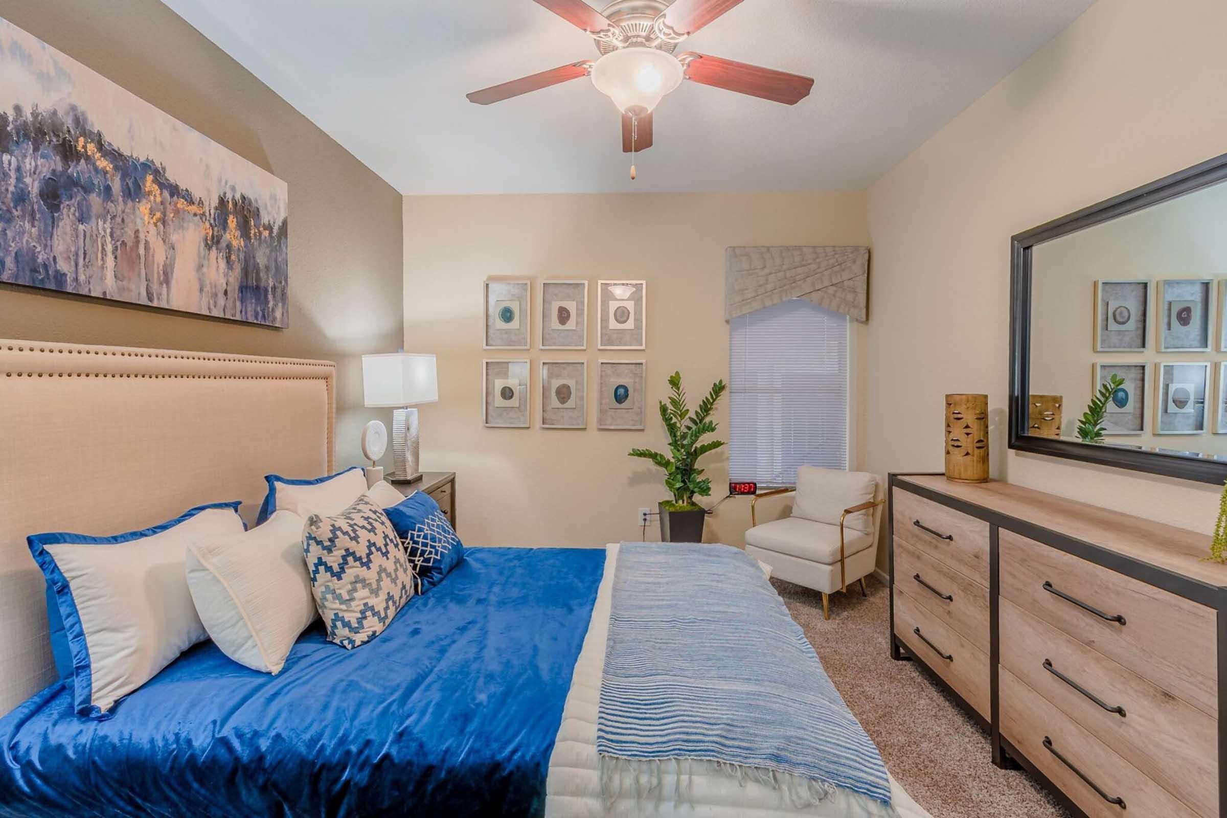 A cozy bedroom featuring a queen-sized bed with blue and white bedding, a wooden dresser, and decorative wall art. A ceiling fan hangs overhead, and there is a comfortable chair in the corner. The room has warm tones and a small potted plant adds a touch of greenery.