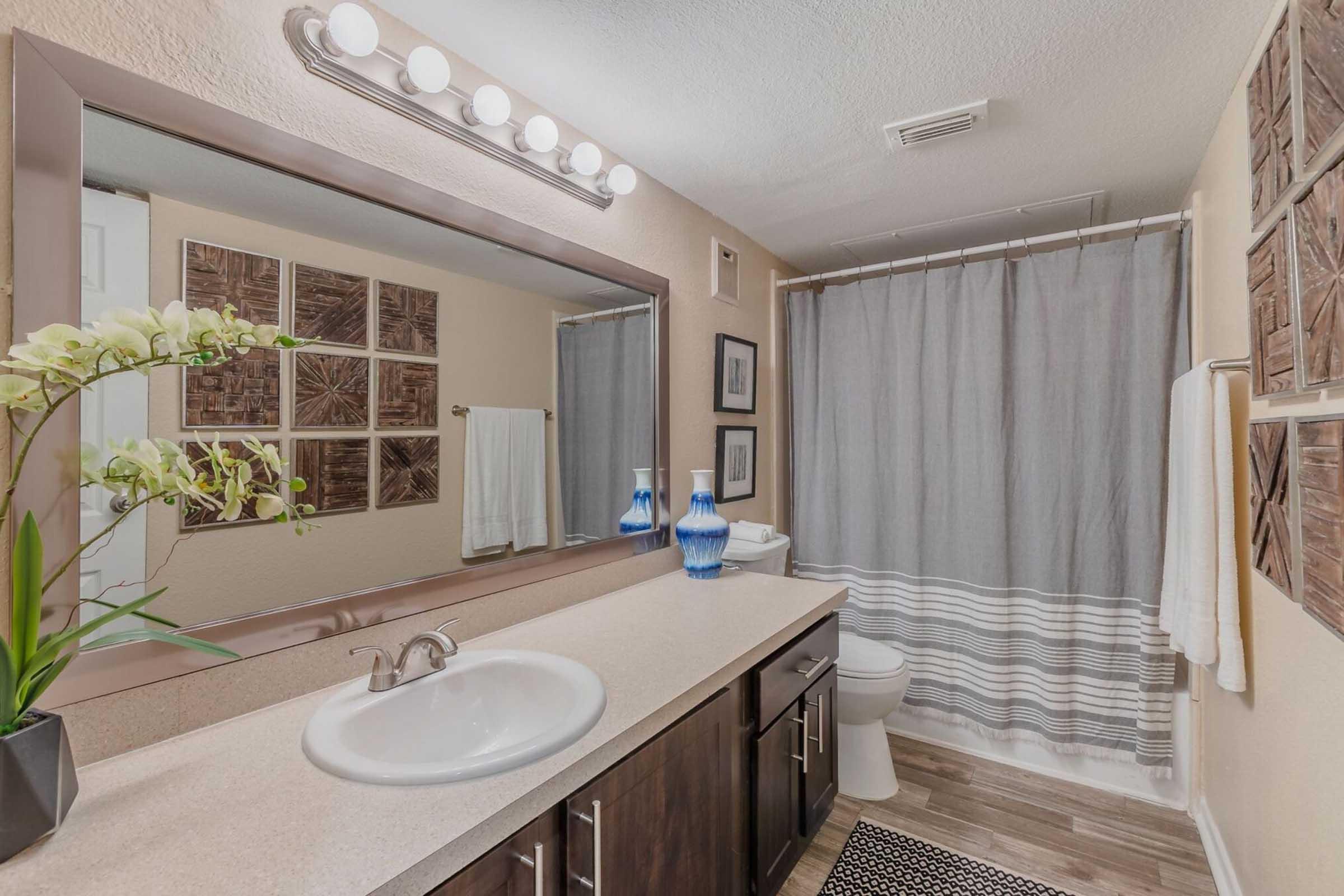 A modern bathroom featuring a large mirror above a dark wood vanity with a sink, topped with decorative items. A light gray shower curtain hangs beside the tub, and a framed wall art display is visible. White towels are neatly arranged, and a plant adds a touch of greenery to the space.