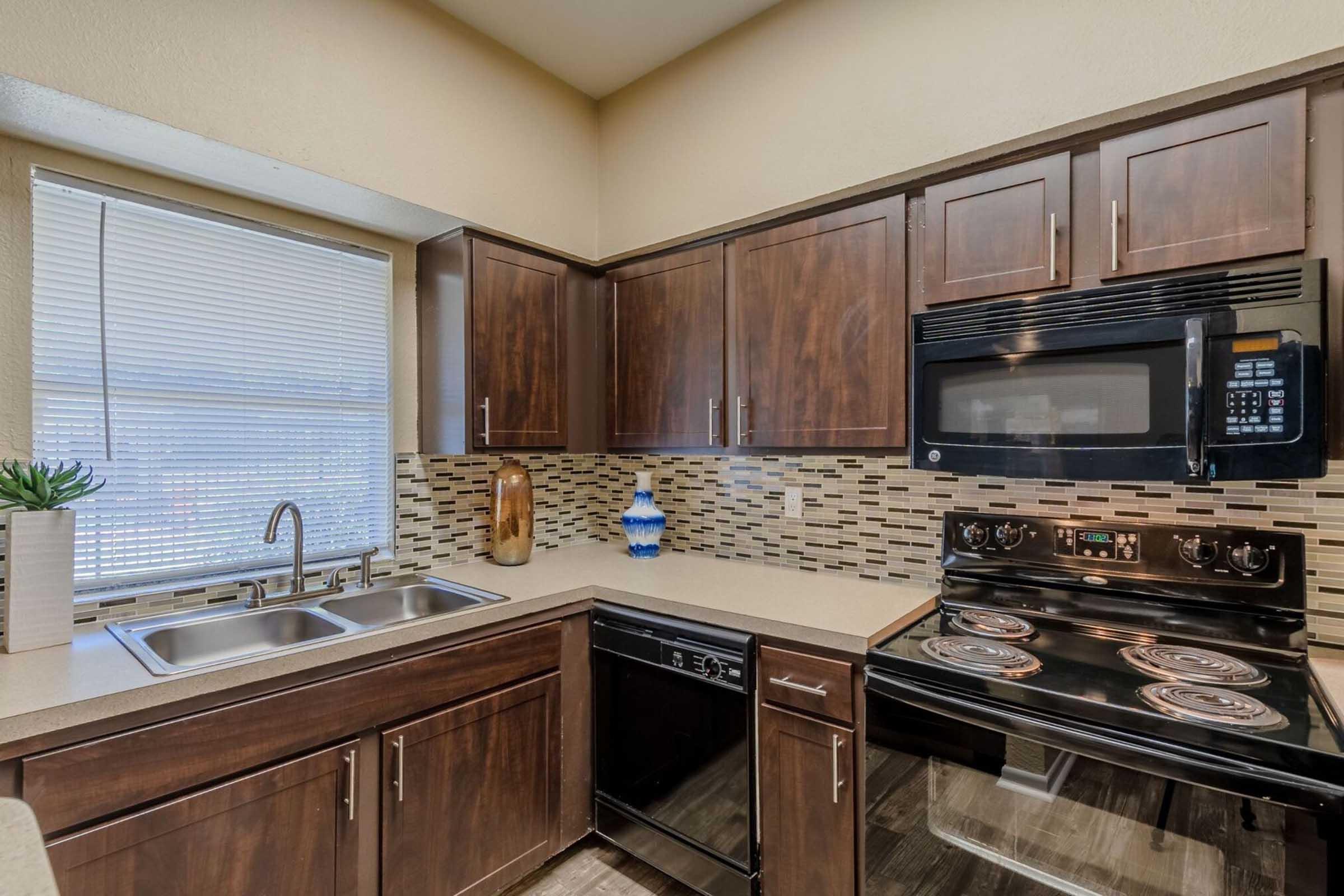 Modern kitchen featuring dark wood cabinetry, a double sink under a window, and a black stove and microwave. The backsplash is made of decorative tiles, and a decorative vase is placed on the countertop. The overall design is sleek and contemporary, with an emphasis on functionality.