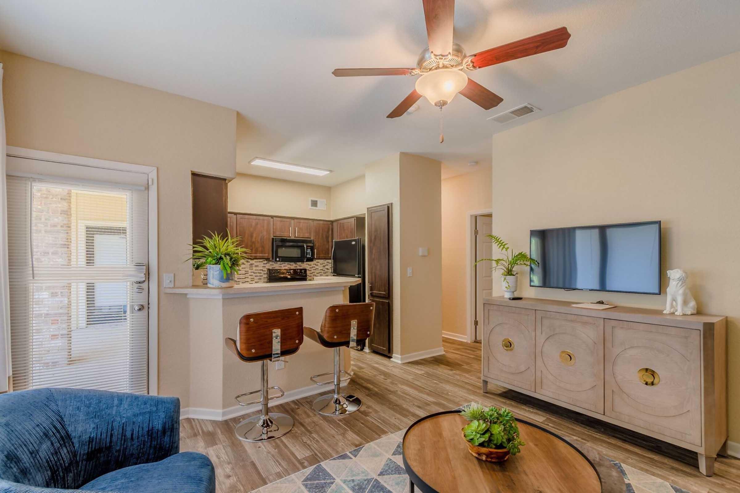A modern, bright living space featuring a blue armchair, a wooden cabinet with decorative items, and a flat-screen TV. The kitchen area has dark cabinetry and stainless steel appliances. Stylish barstools are positioned at a counter. A ceiling fan and natural light enhance the inviting atmosphere.