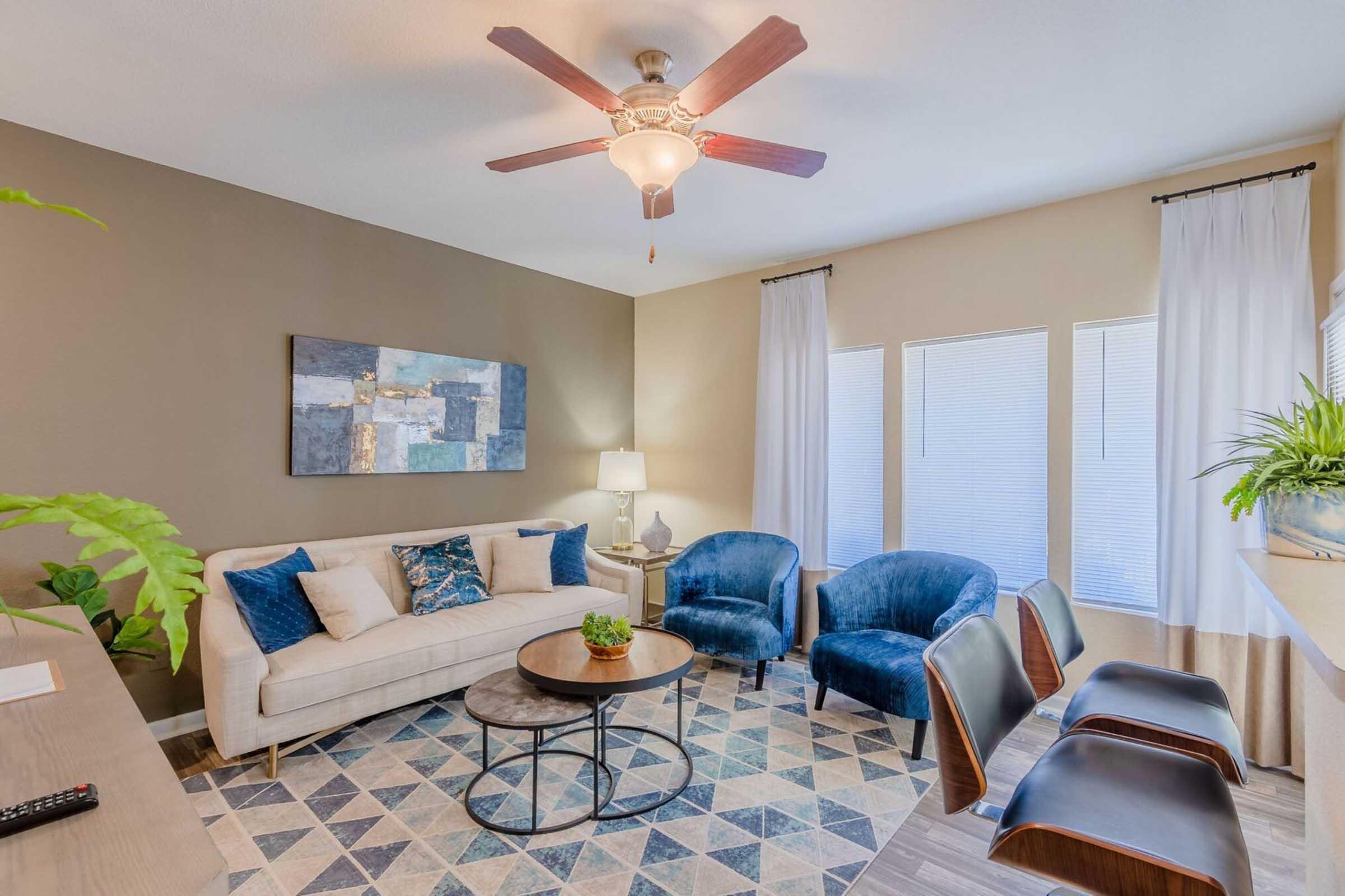 A modern living room featuring a beige sofa with blue and white cushions, two blue armchairs, and a round coffee table. There's a ceiling fan and large windows with sheer curtains. A small plant and decorative artwork are included. The overall design is bright and inviting, with a geometric patterned rug.