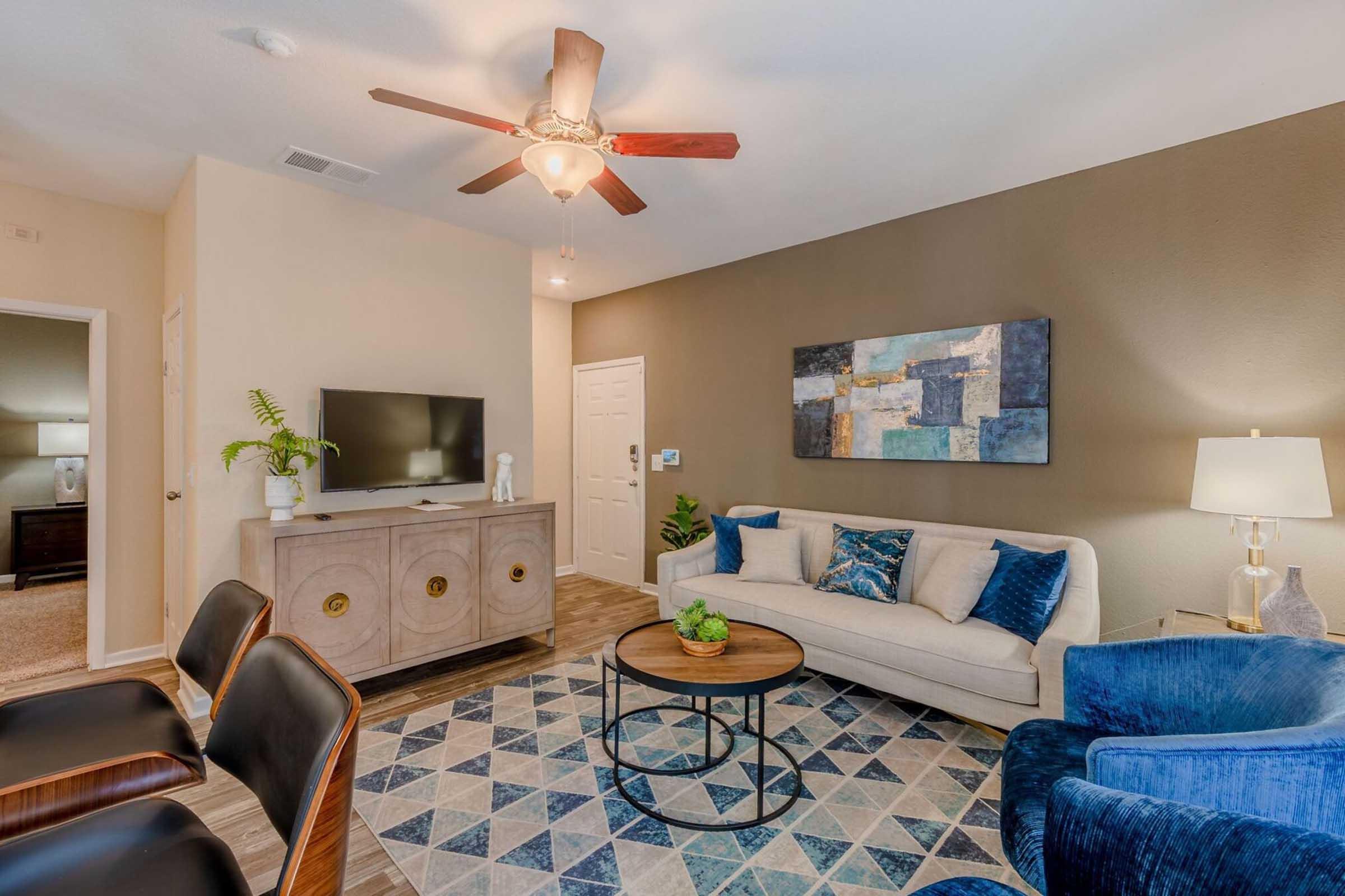 A modern living room featuring a light-colored sofa, a round coffee table with a decorative plant, and a blue accent chair. The room has a ceiling fan, a television on a stylish cabinet, and a wall art piece. The floor is adorned with a patterned area rug, creating a cozy and inviting atmosphere.