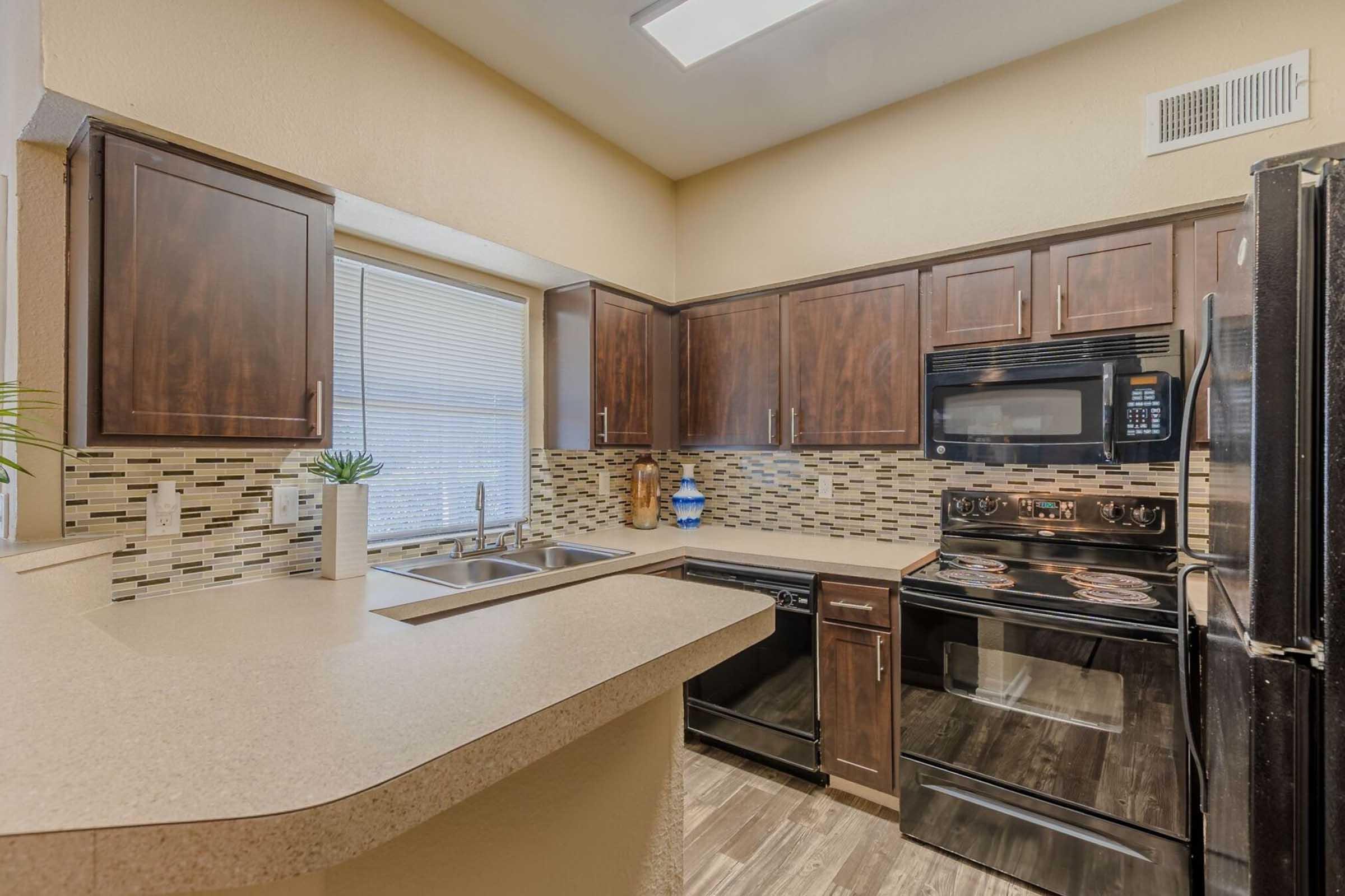 Modern kitchen featuring dark wooden cabinets, a built-in stove, black appliances including a microwave and dishwasher, and a tiled backsplash. A small window provides natural light, and a countertop separates the kitchen from a dining area, with minimal decorative elements.