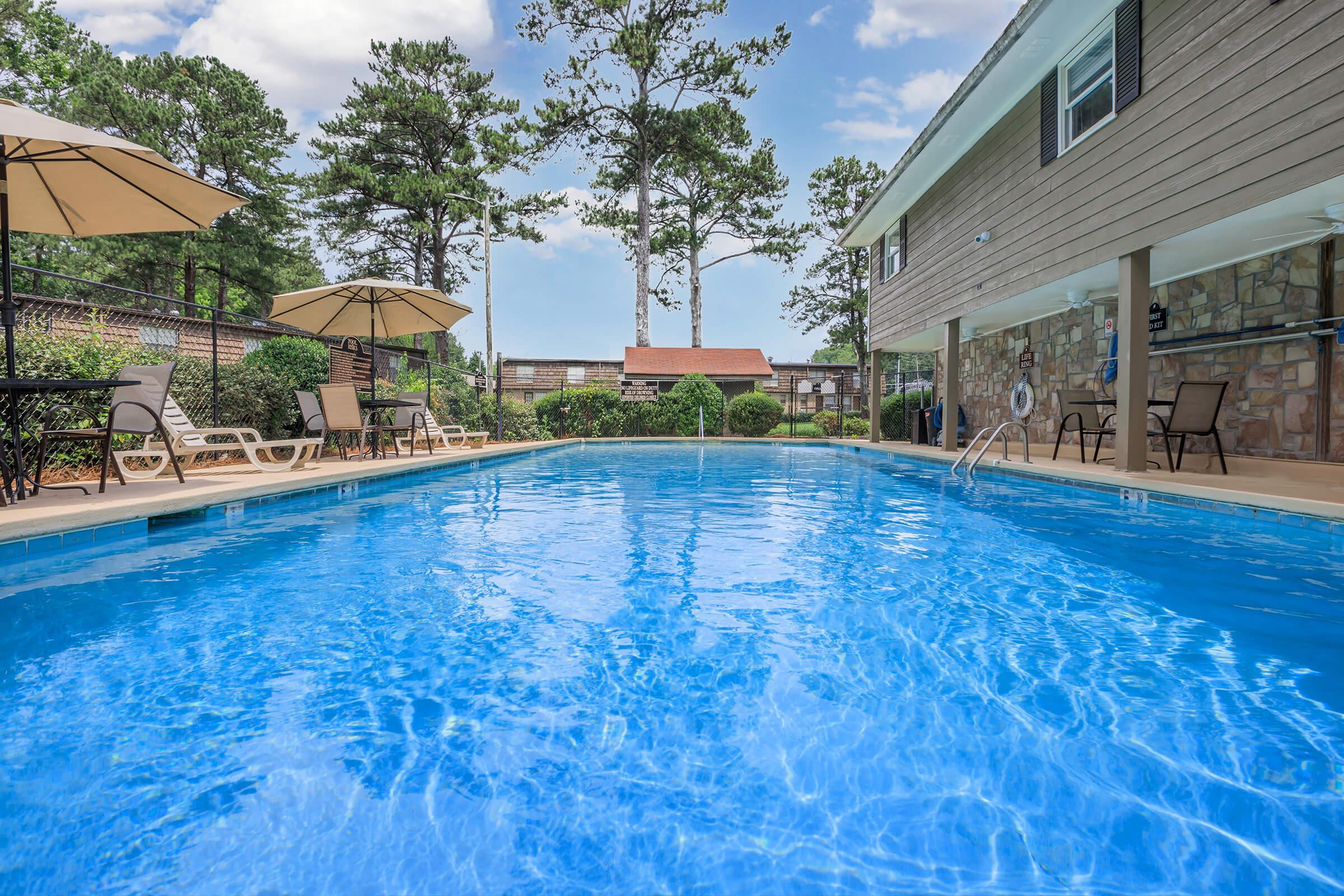A clear blue swimming pool surrounded by lounge chairs and umbrellas. In the background, trees and a house with a stone exterior can be seen. The sky is partly cloudy, creating a serene outdoor atmosphere.