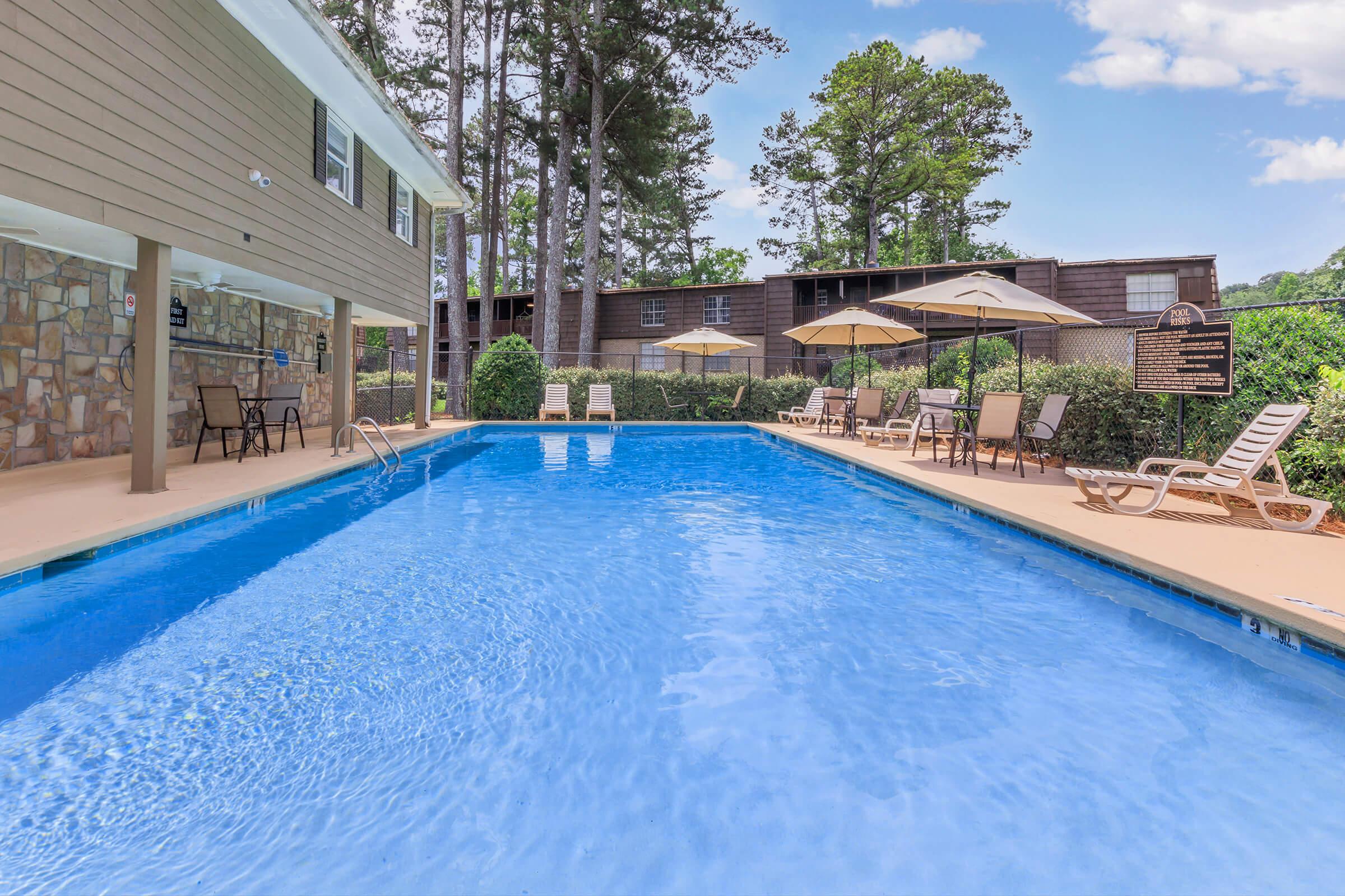 A clear blue swimming pool surrounded by sun loungers and umbrellas, with a backdrop of tall trees and a building. The pool area features a stone wall and several chairs for relaxation, creating a tranquil outdoor space for leisure and enjoyment.