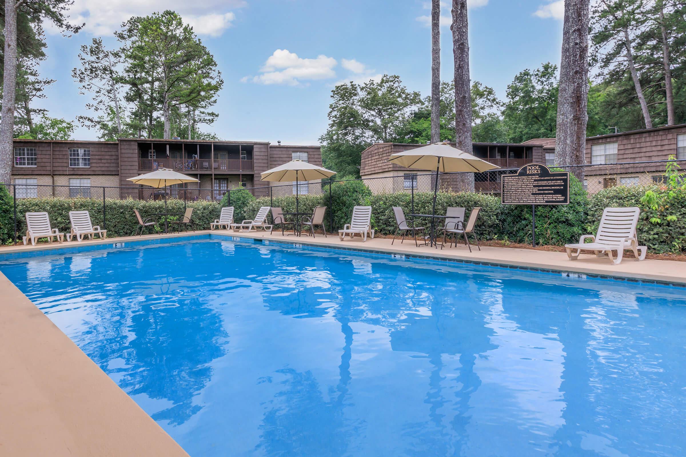A clear blue swimming pool surrounded by chaise lounges and umbrellas, set against a backdrop of greenery and apartment buildings. The scene features tall trees in the background and a sunny sky, creating a relaxing outdoor atmosphere perfect for leisure and recreation.