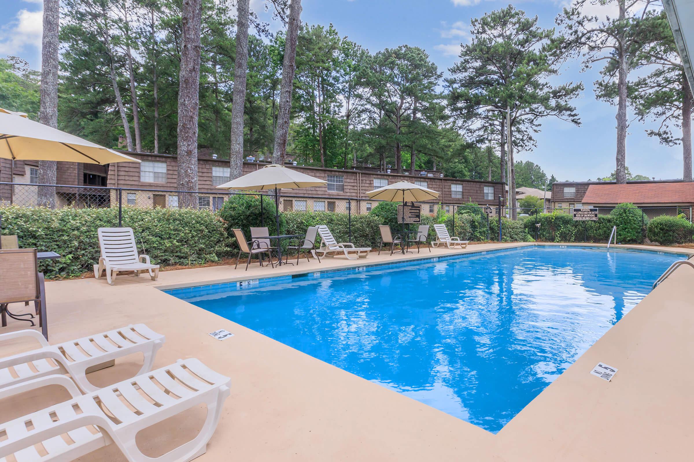 A sparkling blue swimming pool surrounded by lounge chairs and umbrellas, framed by green shrubbery and tall trees. In the background, there are two-story buildings with multiple balconies, creating a relaxed outdoor atmosphere perfect for leisure activities.