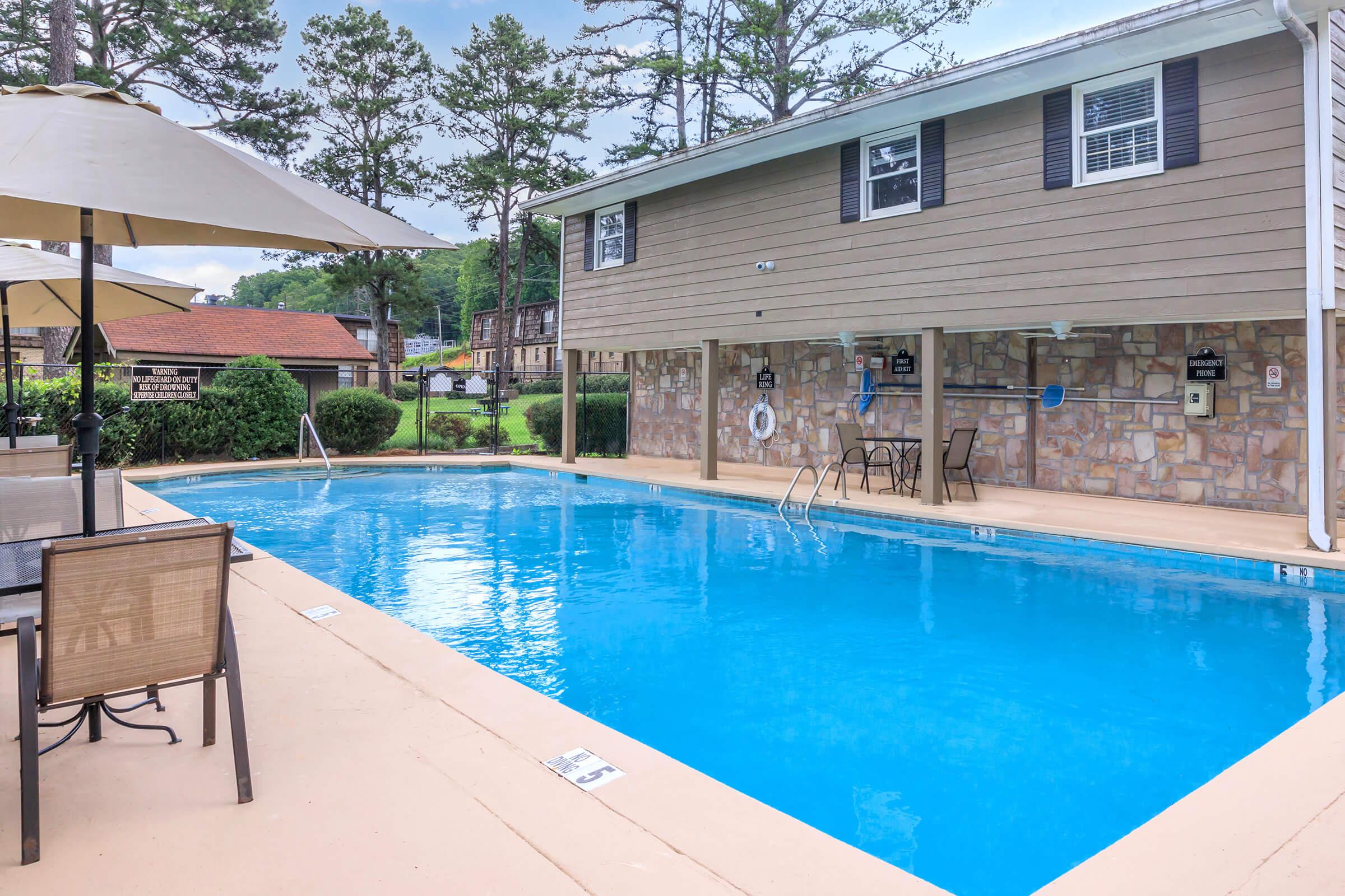 A clear blue swimming pool surrounded by a beige deck. To the left, there are tables and chairs under an umbrella. In the background, a two-story beige building with a stone facade and several trees nearby. The scene conveys a relaxed outdoor atmosphere, ideal for leisure and recreation.