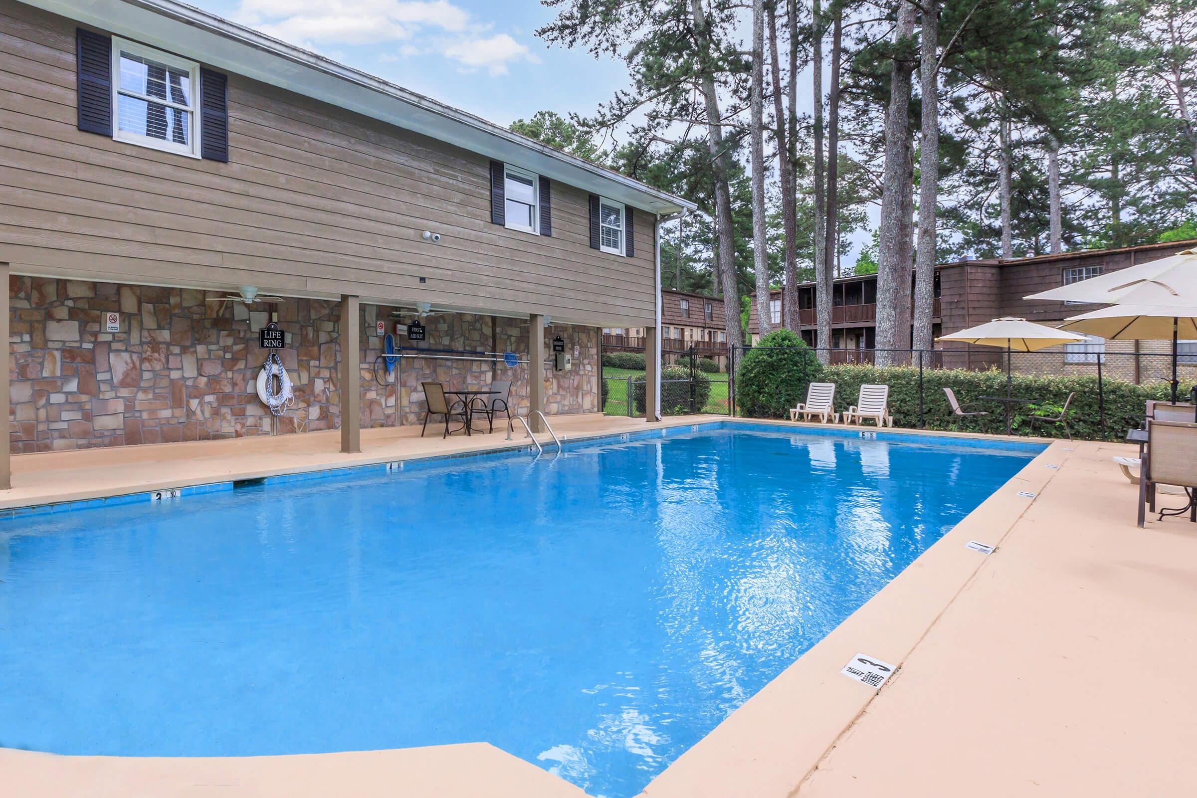 A clear blue swimming pool surrounded by a beige patio, with lounge chairs and umbrellas nearby. In the background, a wooden building with windows and a stone wall can be seen, along with tall trees providing shade. The scene is tranquil and inviting, ideal for relaxation.