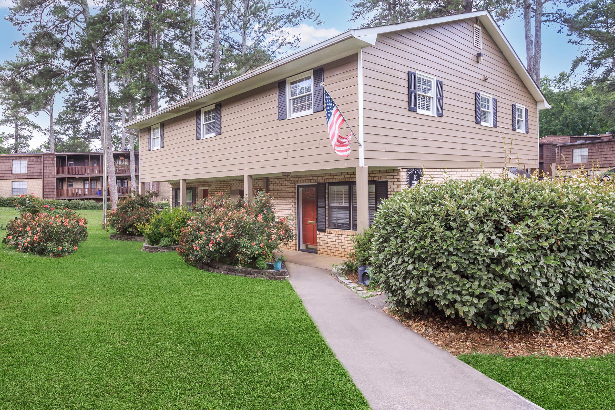A two-story house with a light brown exterior, featuring a red door and white trim. The front yard has well-maintained green grass and flower bushes. An American flag is displayed on the porch. Surrounding trees and a walkway lead to the entrance, giving a welcoming suburban feel.