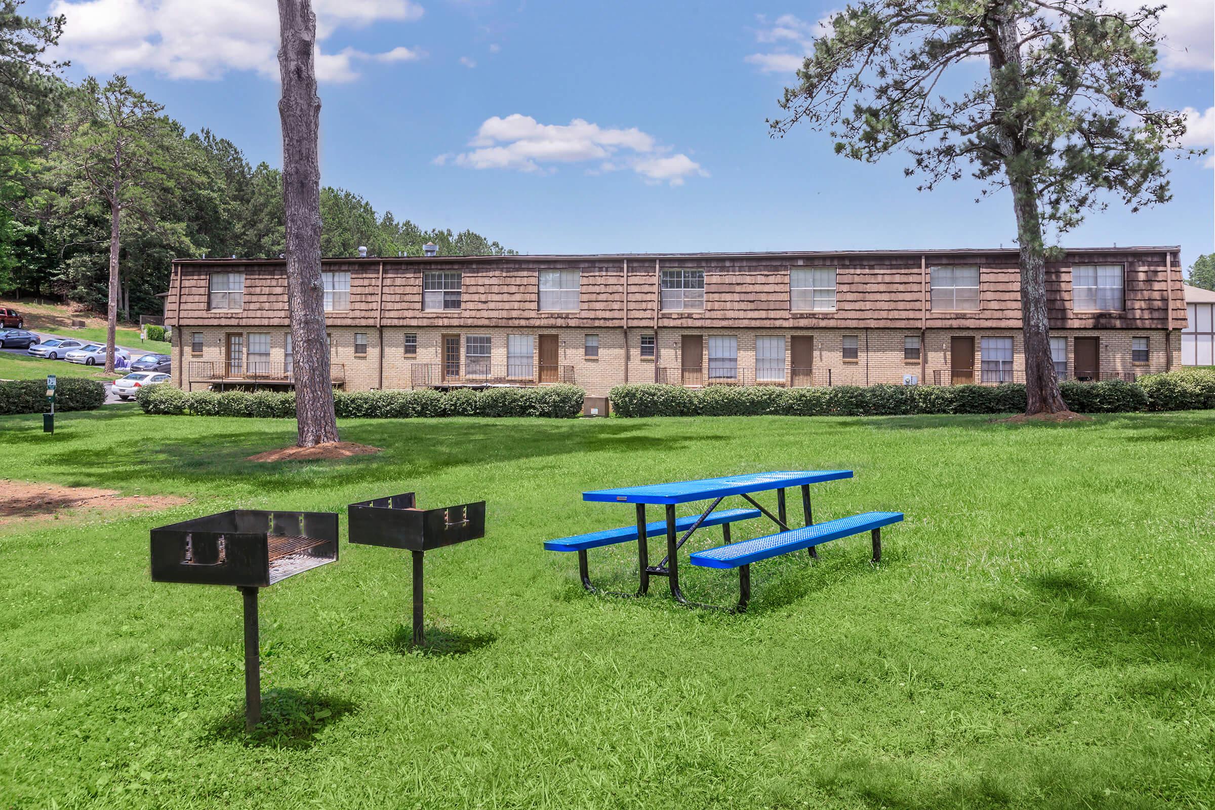 A grassy area with a blue picnic table and two charcoal grills in front of a brick apartment building. The background features trees and a blue sky with scattered clouds.