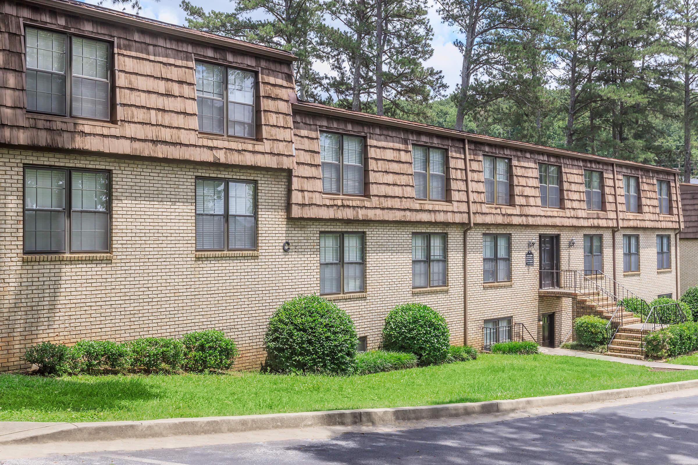 A two-story brick building with brown shingle roofing, featuring several large windows and a set of stairs leading up to the entrance. The surrounding area has neatly trimmed green shrubs and grass, set against a backdrop of tall trees under a clear sky.