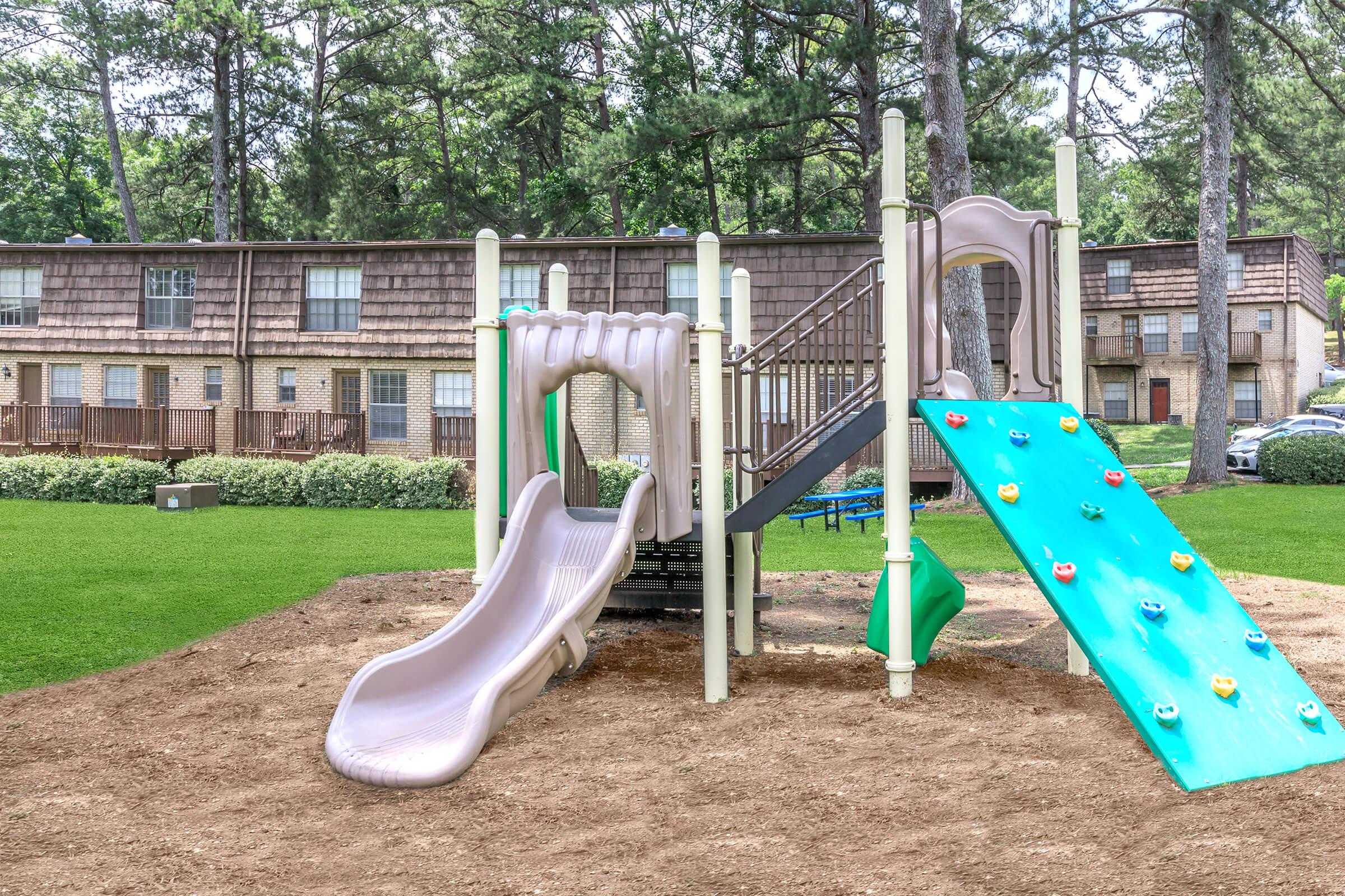 A children's playground featuring a slide, climbing wall, and steps. It is set on sandy ground surrounded by grass, with several trees in the background. In the distance, there are residential buildings partially visible.