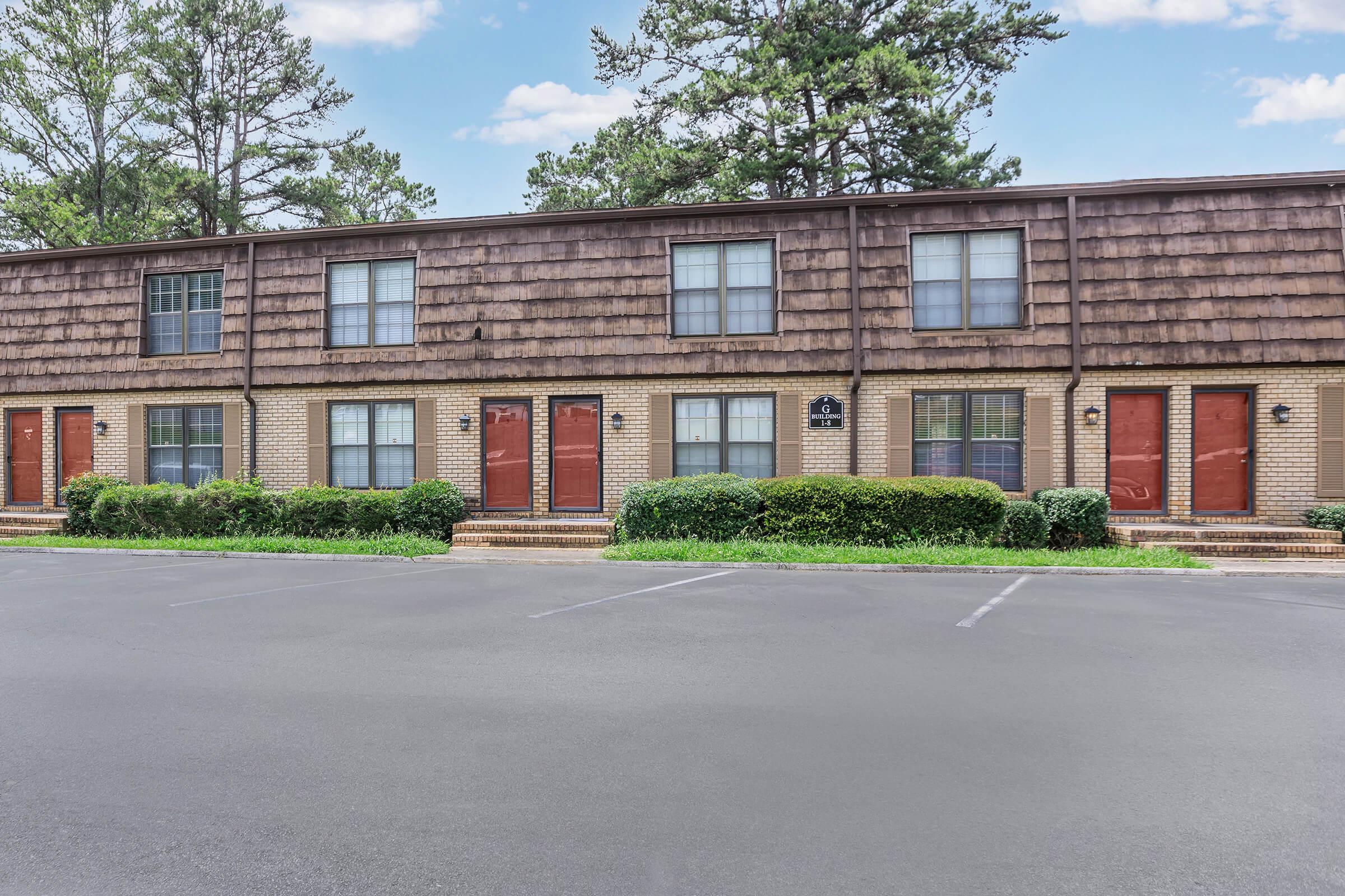 A row of two-story townhouses with brown shingles on the upper level and brick on the lower level. Each unit has large windows and a red door, with neatly trimmed hedges in front. A paved parking area is visible, with several parking spaces marked. The scene is set against a backdrop of trees and a blue sky.