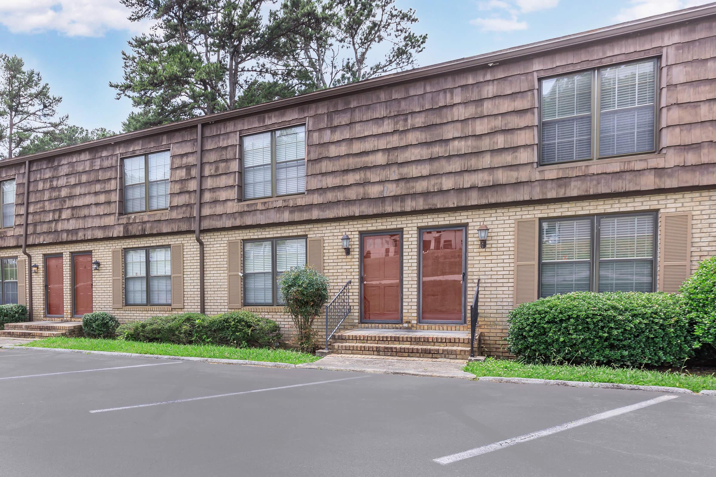 A row of townhouses with brown shingle siding and brick bases. Each unit features a front door, windows, and shrubs along the walkways. The parking lot in front is partially visible, with a few empty parking spaces. Trees are in the background under a partly cloudy sky.