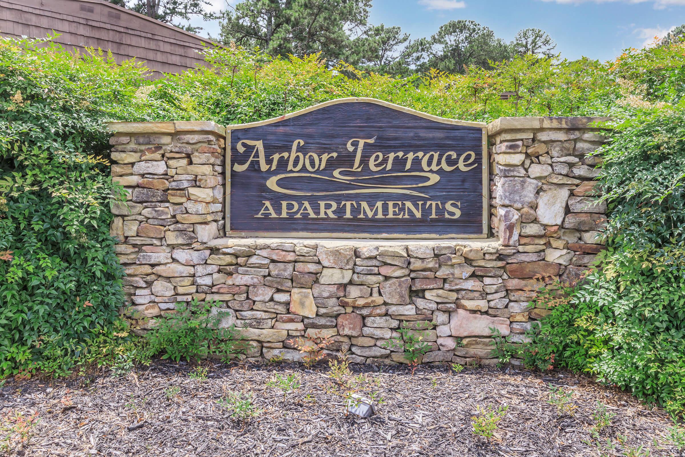 Wooden sign for "Arbor Terrace Apartments" displayed on a stone wall, surrounded by greenery. The sign features elegant lettering and a decorative design. The background includes a blue sky with some clouds and trees, creating a serene atmosphere.