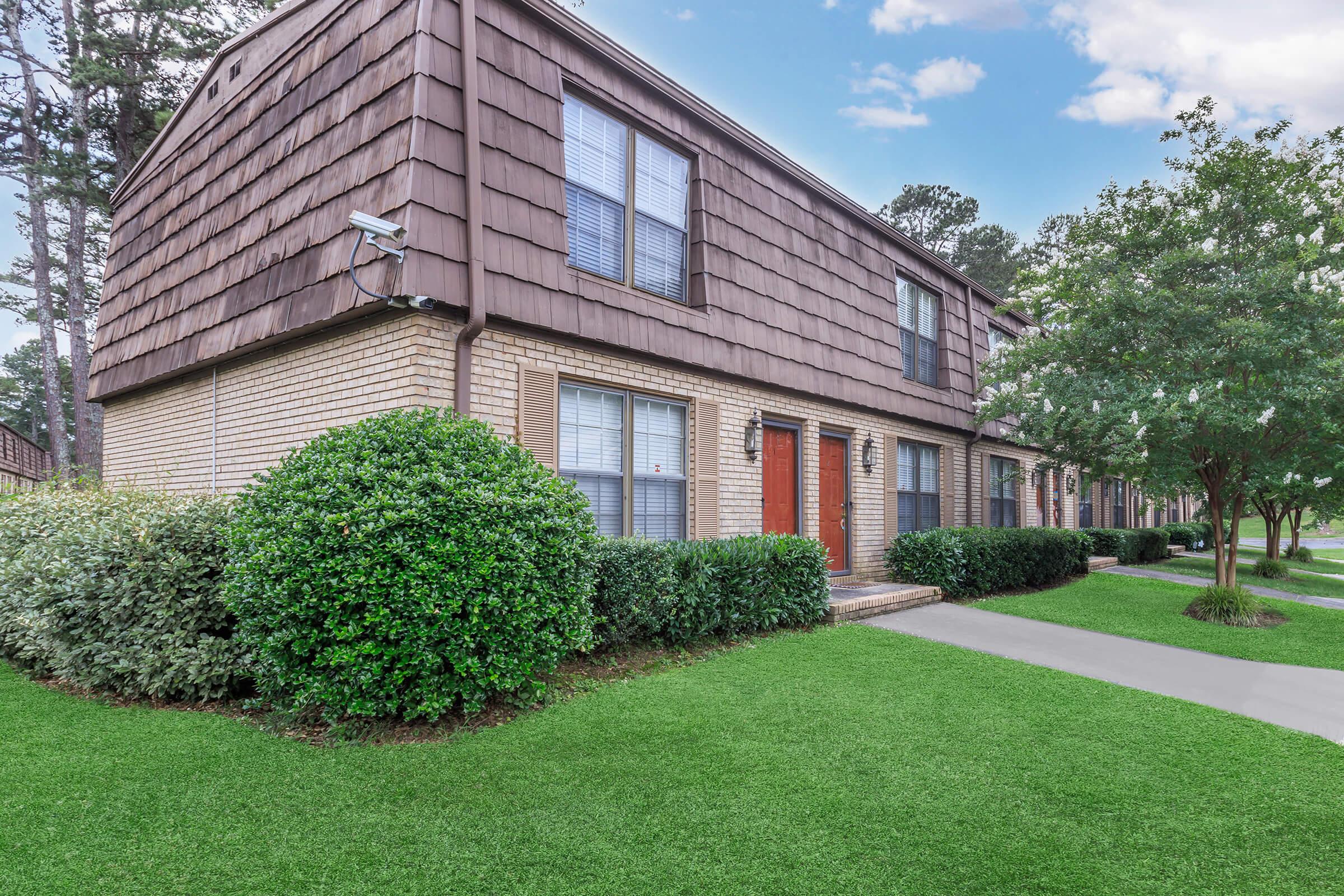A well-maintained two-story apartment building with a brown shingle roof, featuring several windows and two entrance doors. Lush green shrubs and neatly trimmed grass frame the building, while a concrete pathway leads to the entrances. The background includes trees under a clear sky.