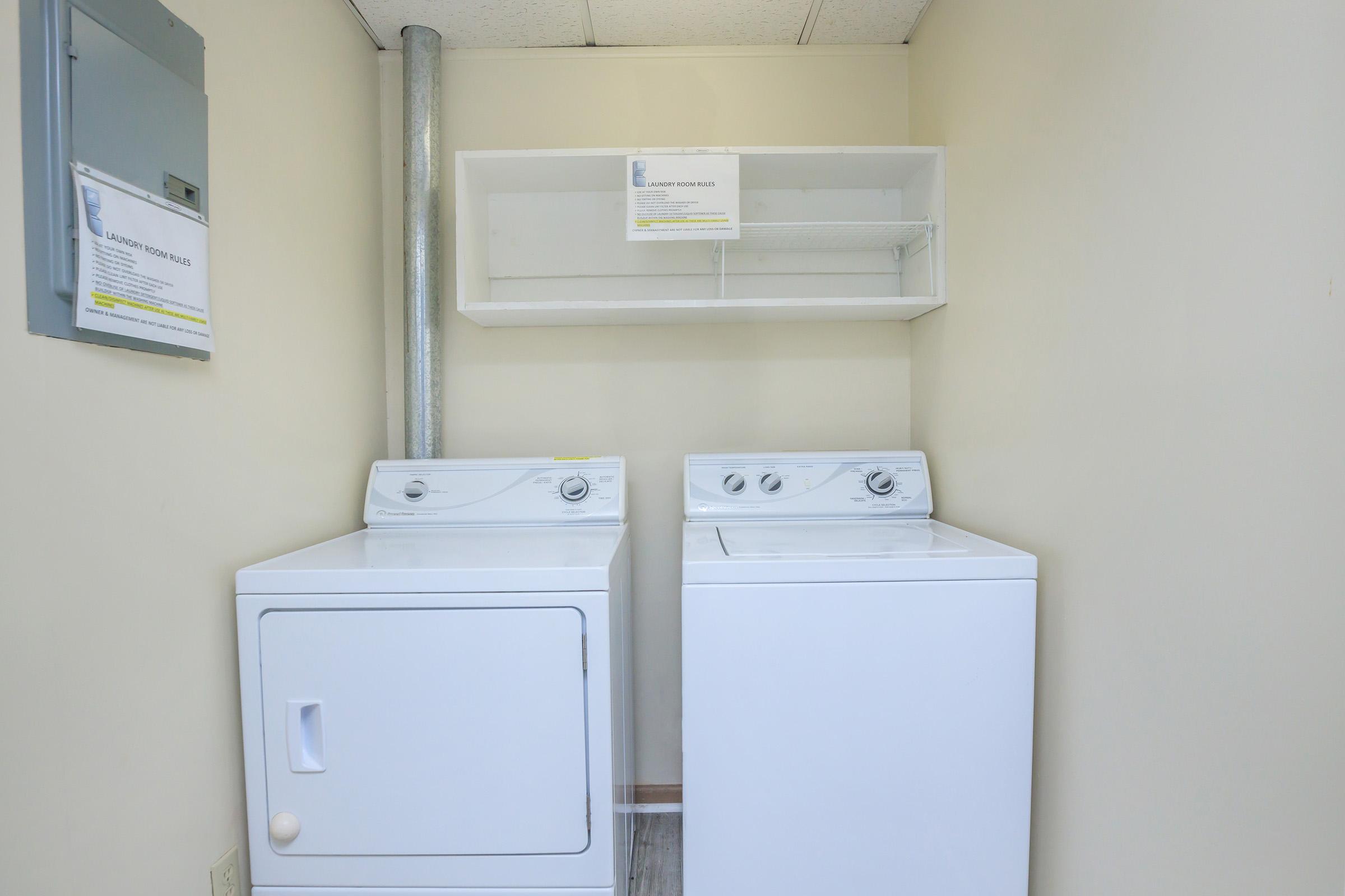 A laundry room featuring a white washing machine and a dryer side by side, with a shelf above them. The walls are a light color, and there's an electrical panel on the far left. A sign is placed on the shelf with laundry information. The floor appears to be a light wood or laminate material.