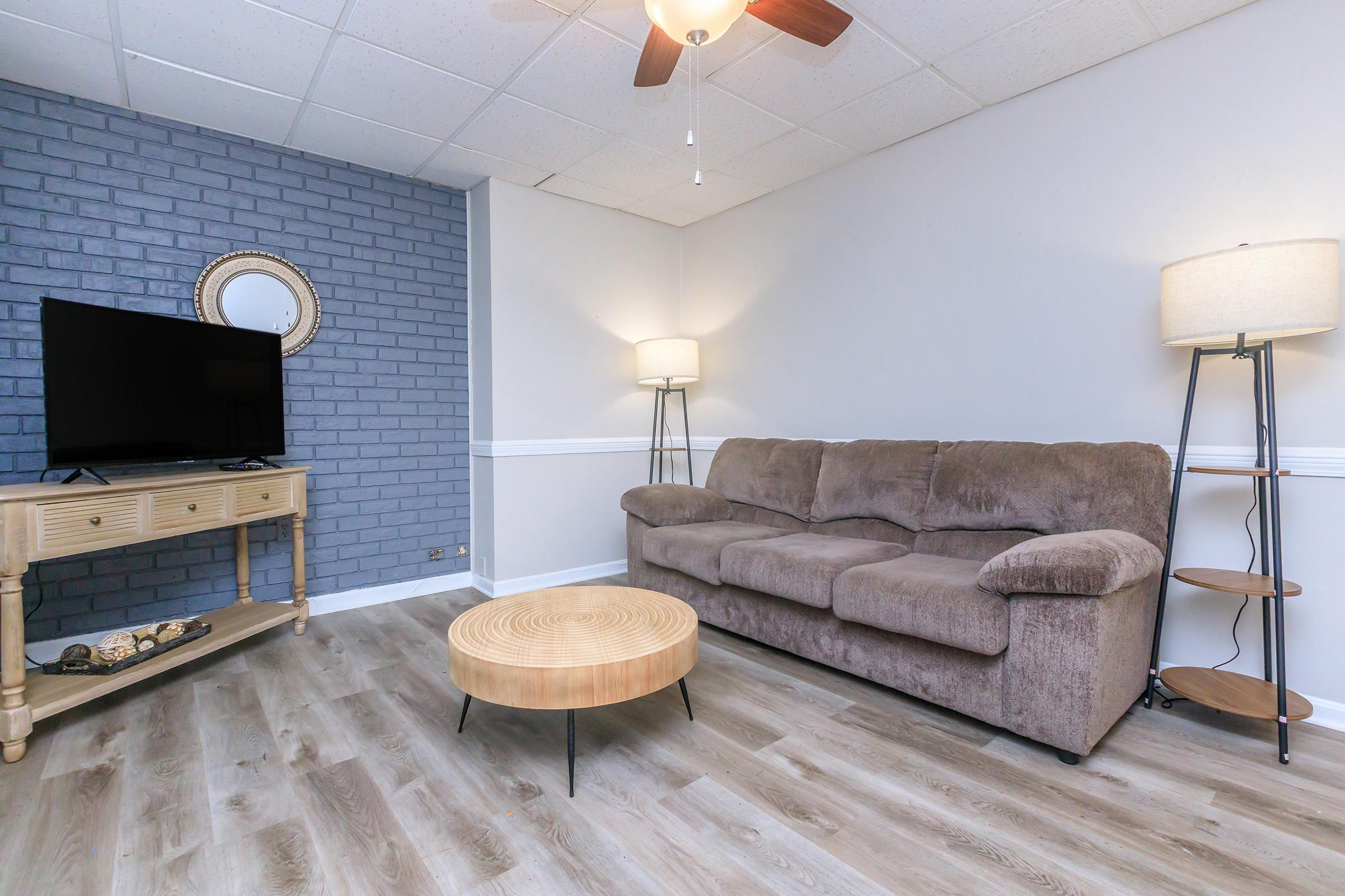 Cozy living room featuring a brown sofa, a wooden coffee table, a TV on a light wood console, and a standing lamp. The walls are painted in light tones, with one wall showcasing gray brick. Natural light fills the space, creating a warm and inviting atmosphere.