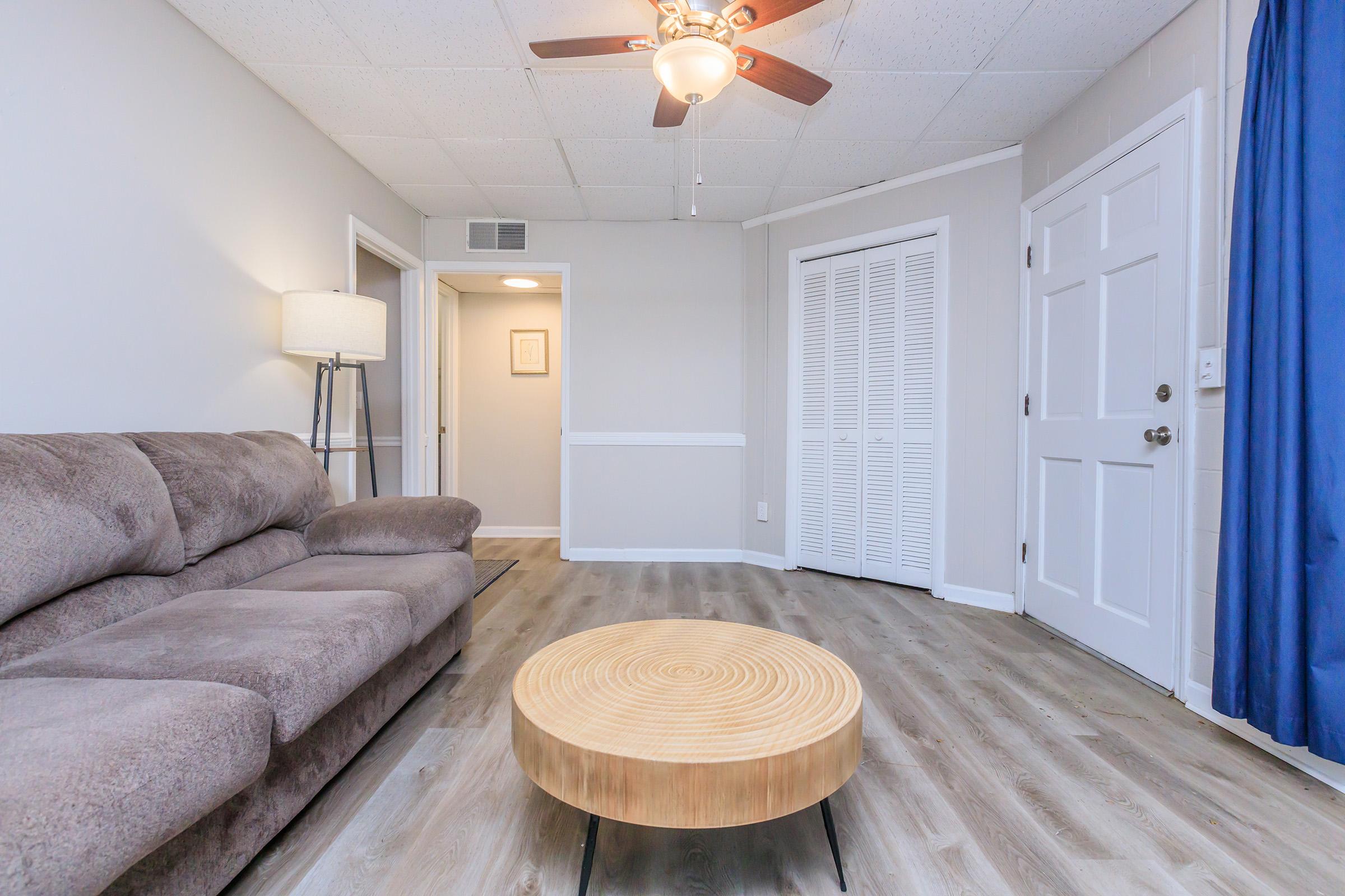 A cozy living room featuring a brown sectional sofa, a circular wooden coffee table, and a ceiling fan. The room has light gray walls, a blue curtain, and a closet with white bi-fold doors. A doorway leads to another room, creating a warm and inviting atmosphere.
