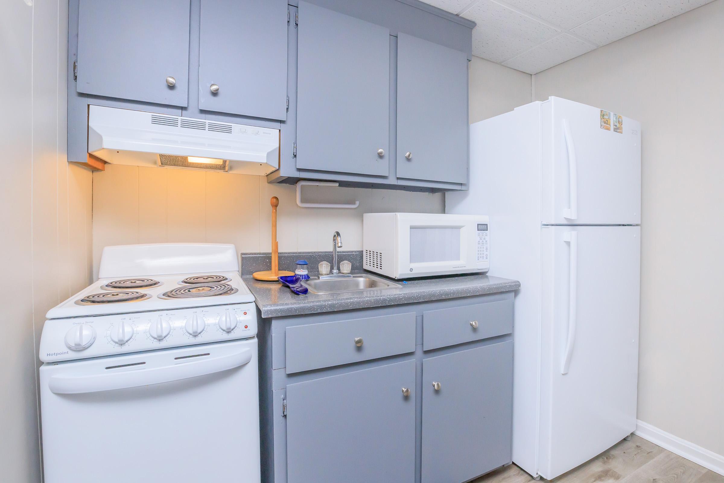 A small kitchen scene featuring gray cabinets, a white refrigerator, and a white microwave. There is a stove with four burners, a sink, and a countertop with a cutting board and a bottle of water. The walls are a light beige color, creating a cozy and functional space.