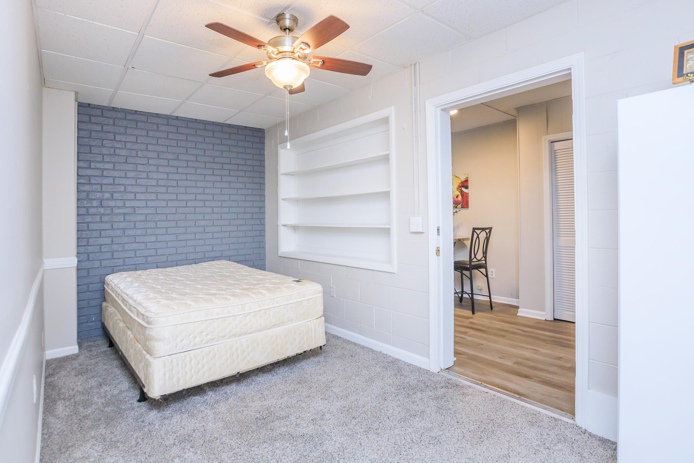 A neatly arranged bedroom featuring a light-colored mattress on a base, a ceiling fan, and a textured gray accent wall. Shelves are built into the wall, and a doorway leads to another room, visible in the background. The floor is carpeted, creating a cozy atmosphere.