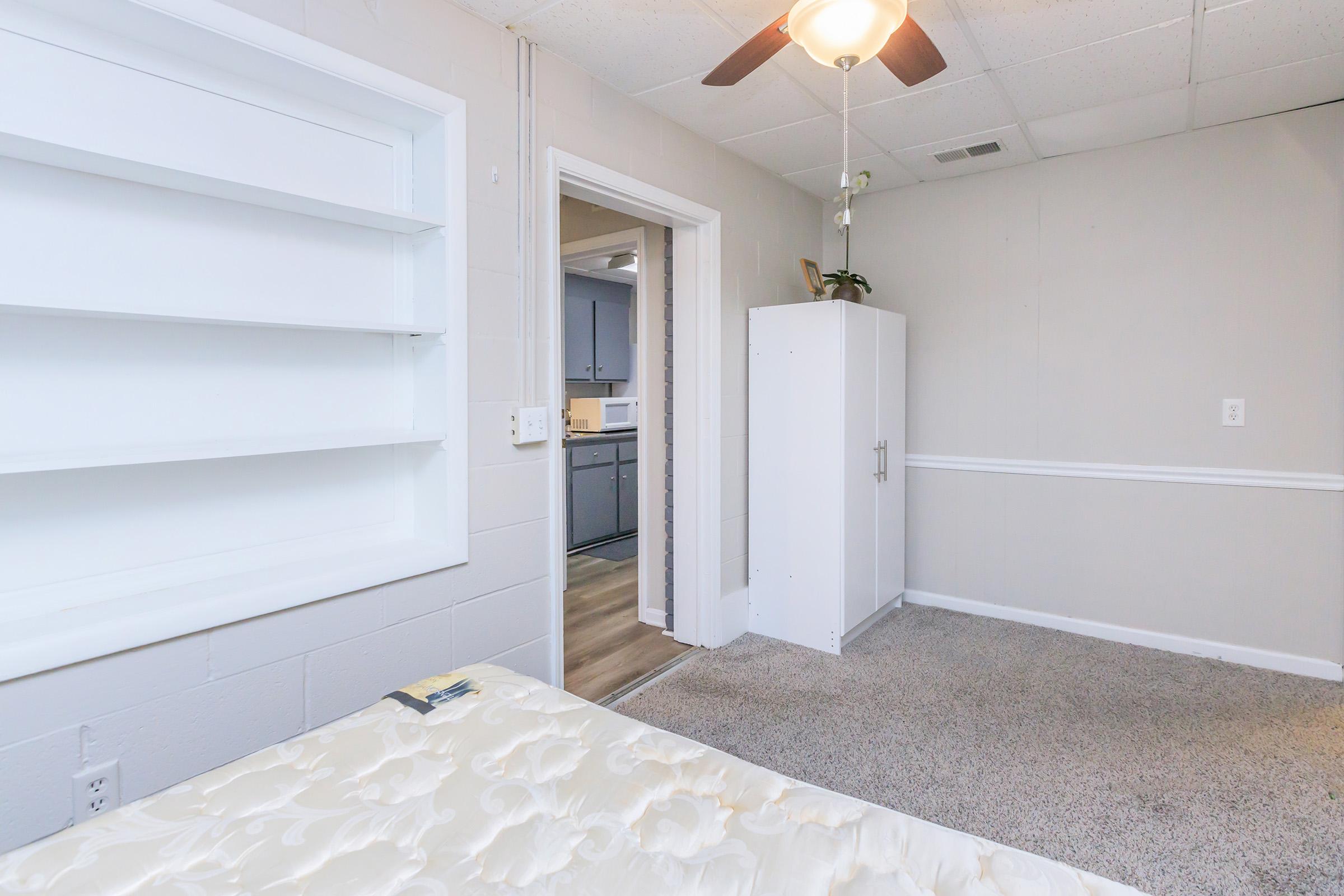 A room with light-colored walls and carpeted flooring. It features a simple ceiling fan and a white, built-in shelf unit. There is a door leading to another room and a white cabinet in the corner. A mattress is placed on the floor, suggesting the room is in the process of being furnished or set up.