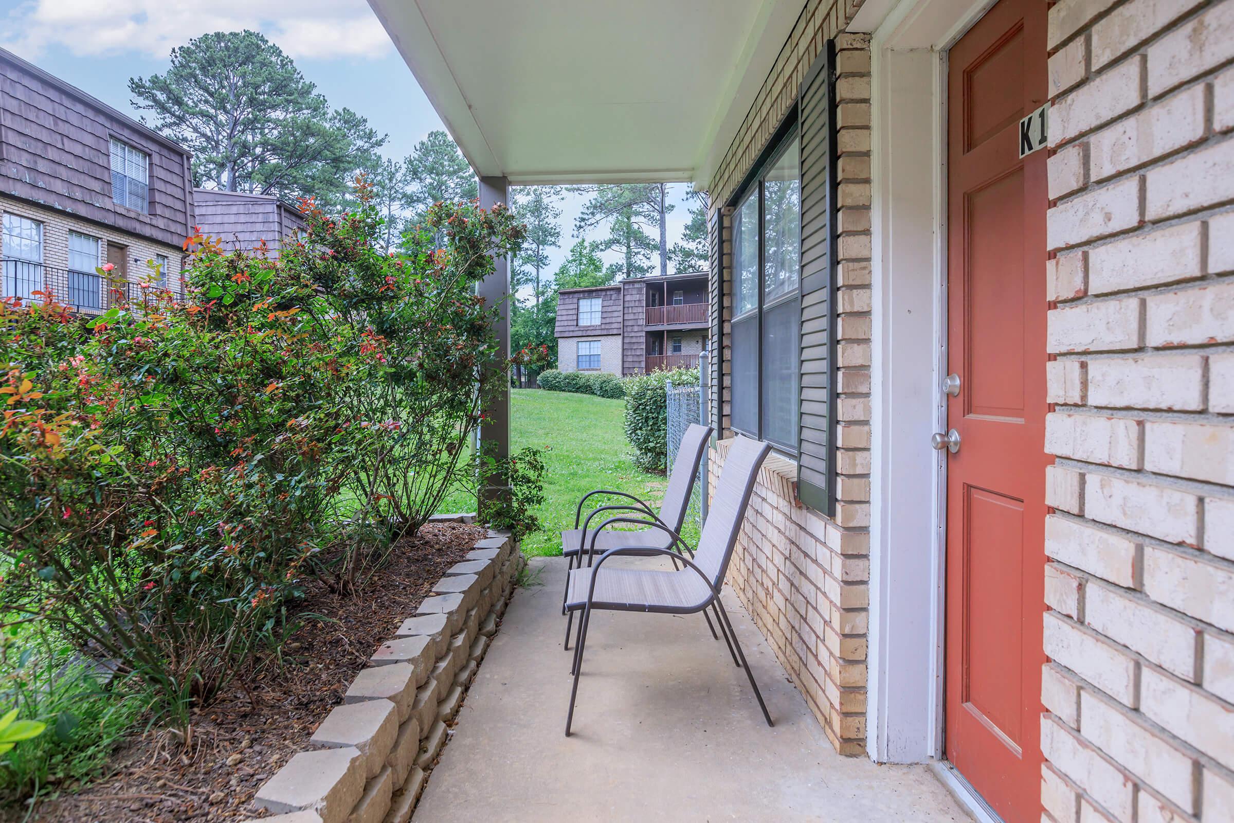A small porch area in front of an apartment, featuring two outdoor chairs and a brick pathway. Lush greenery and bushes with vibrant flowers surround the entrance. The door is painted orange, and there are windows that overlook the lawn and nearby buildings.