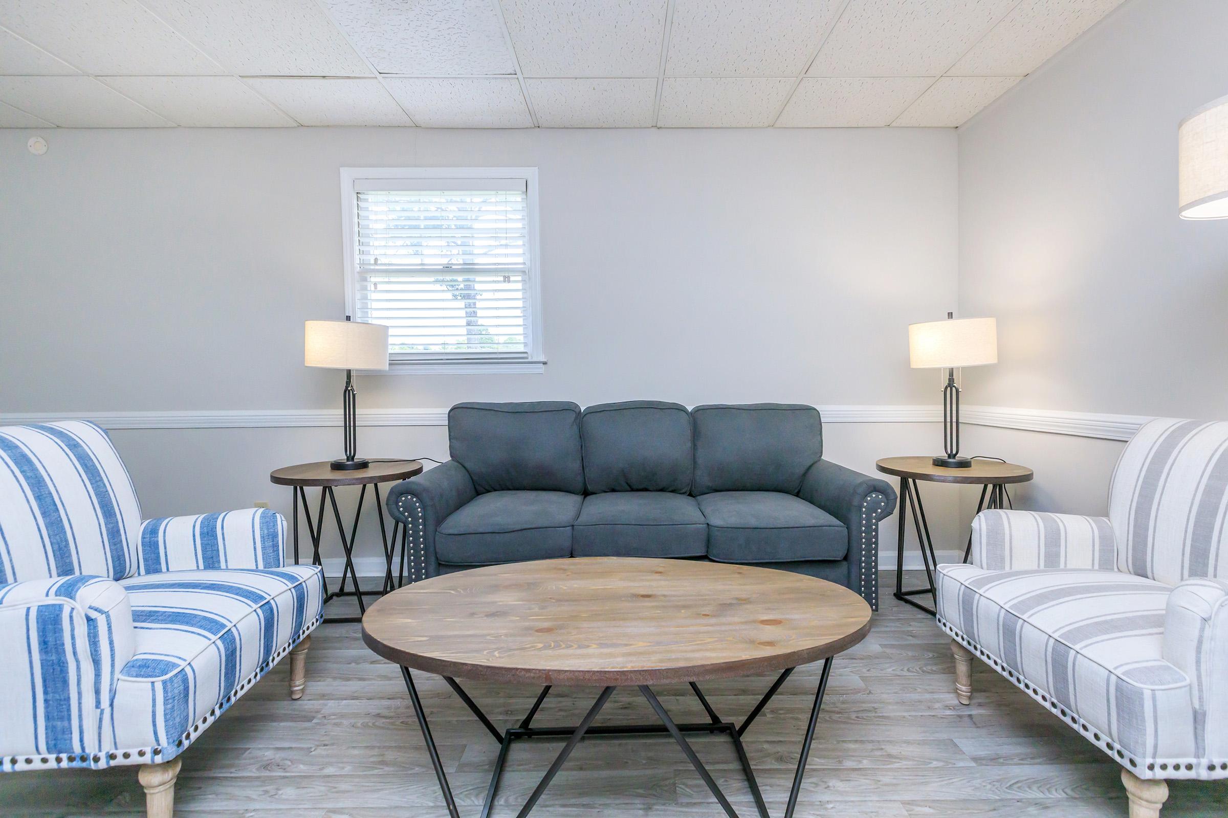 A cozy living room featuring a gray couch in the center, flanked by two armchairs with blue and white stripes. A wooden coffee table sits in front, with two side tables holding lamps. The walls are light gray, and natural light comes through a window with white blinds.
