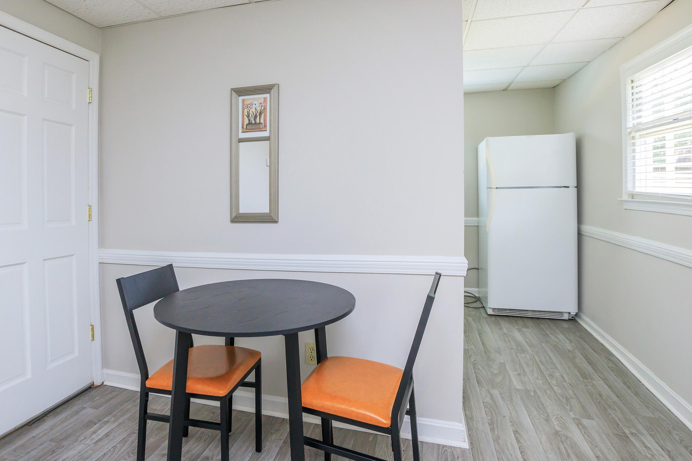 A small dining area featuring a round black table with two chairs, one orange cushion, and a framed picture on the wall. In the background, there is a white refrigerator and light-colored walls with wainscoting. The space is well-lit with natural light from a window.