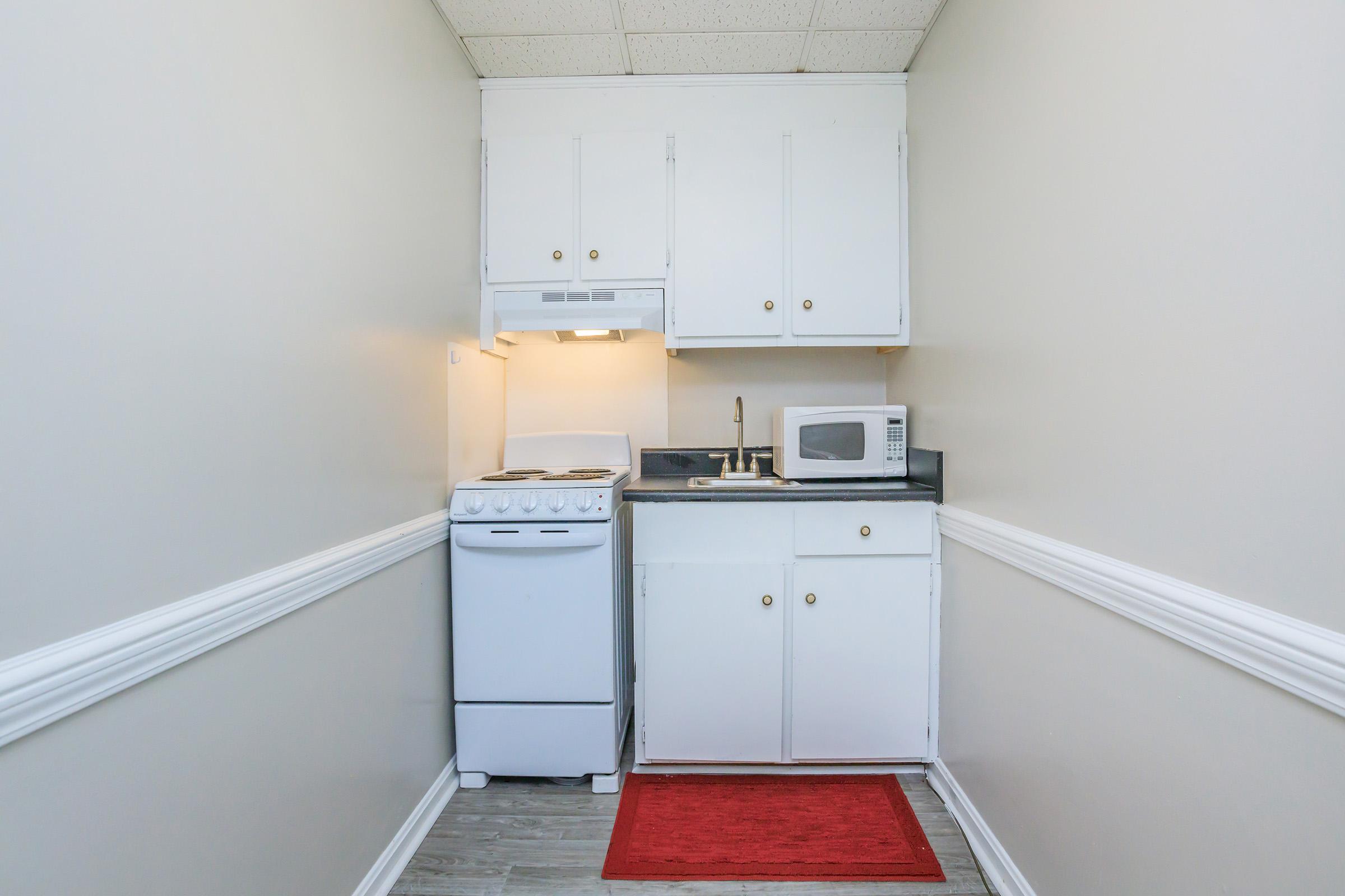 A small kitchen space with white cabinetry, a white stove, a microwave, and a sink. The countertop is dark, and there is a red rug on the floor, creating a cozy atmosphere. The walls are painted a light color, enhancing the bright and clean look of the kitchen area.