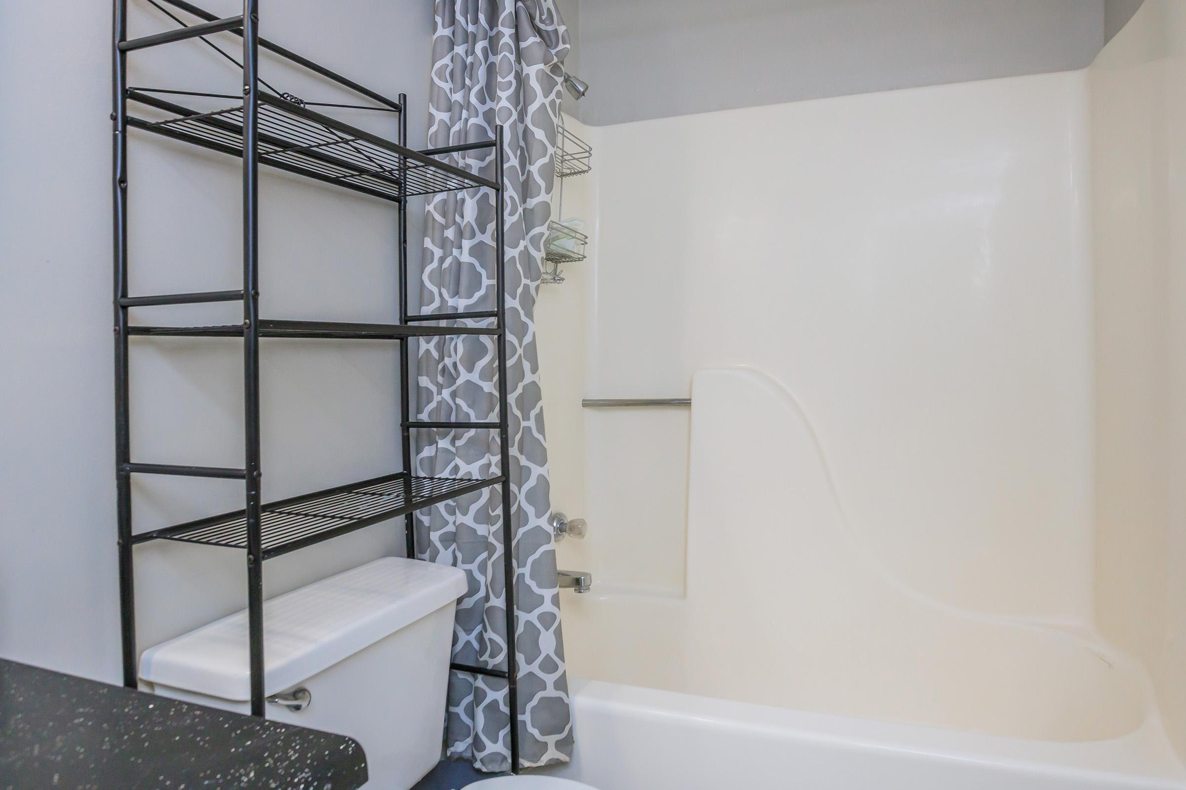 A clean, modern bathroom featuring a white bathtub with a shower, a black metal shelving unit, and a decorative gray and white shower curtain. The walls are painted a light color, and there is a black countertop visible in the foreground.