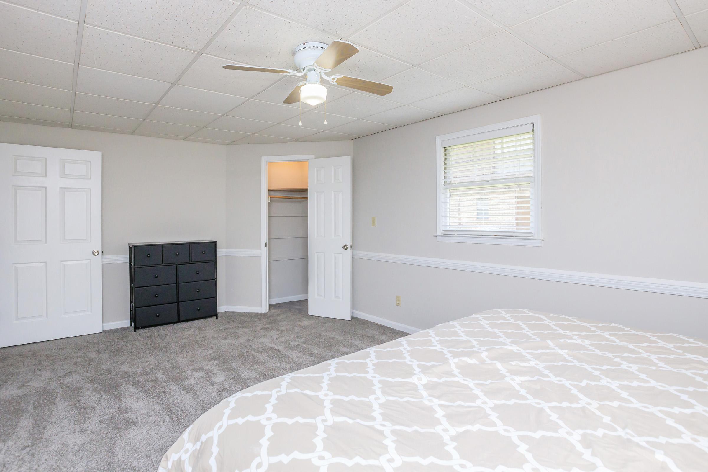 A spacious, well-lit bedroom featuring a ceiling fan, light-colored walls, and a carpeted floor. There is a bed with a patterned duvet in the foreground, a black dresser against the wall, and a closet with double doors. A window allows natural light to enter the room.