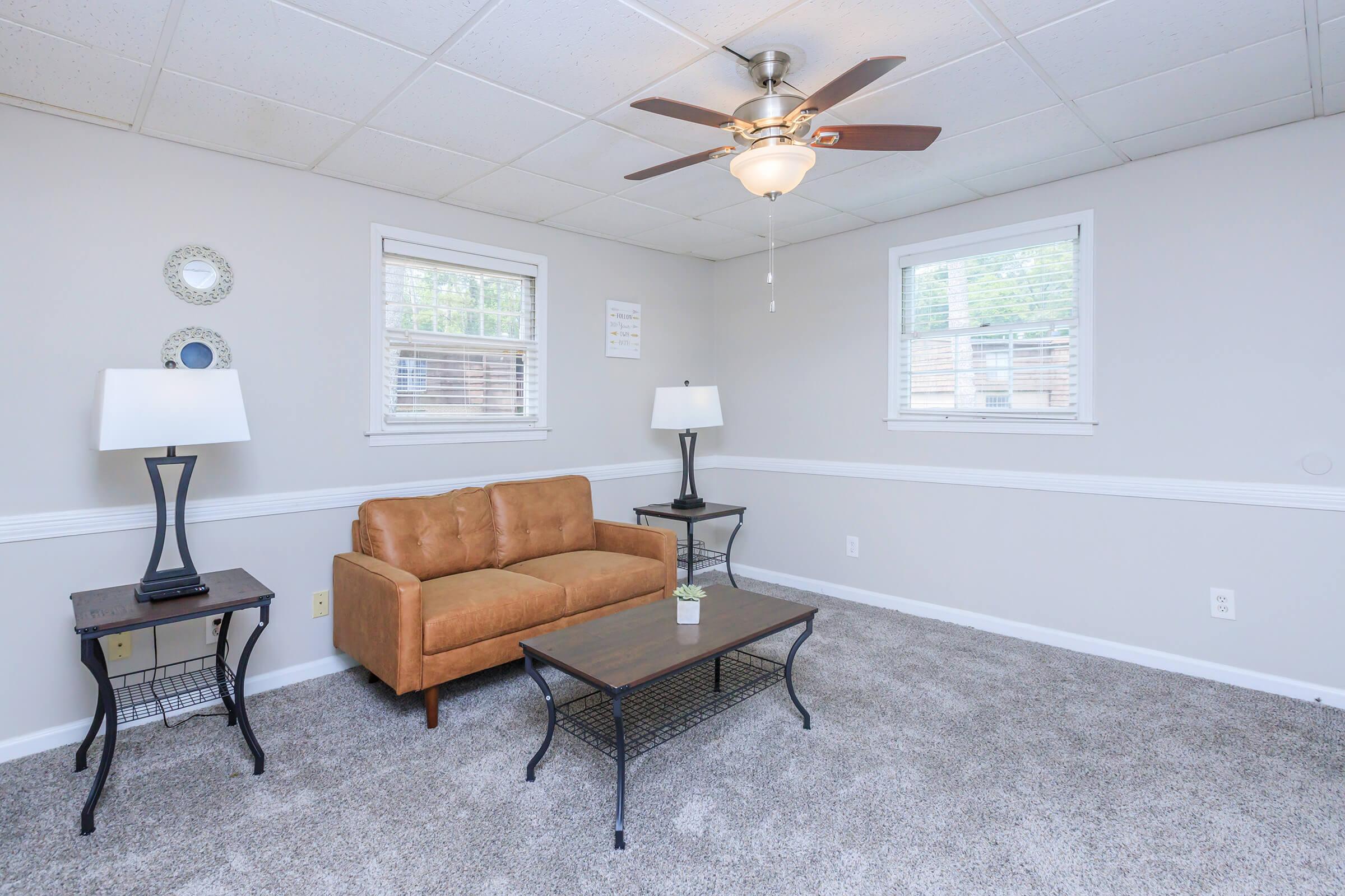 Cozy living room featuring a brown sofa, a black coffee table, and two lamps on side tables. Light gray walls and a ceiling fan complete the space. Natural light streams through two windows, and a small plant decorates the table. Carpet adds warmth to the room.