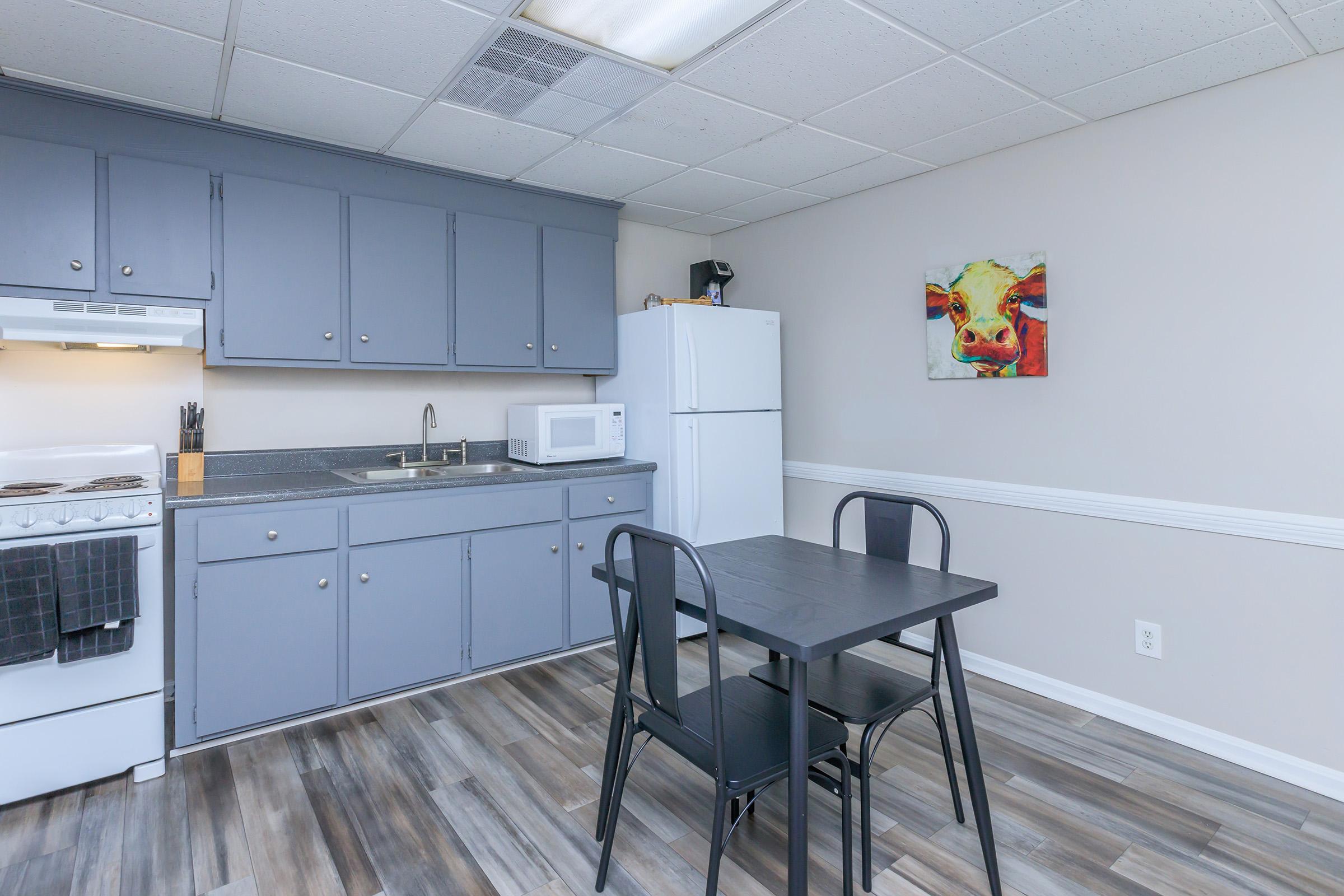 A modern kitchen featuring gray cabinets, a white stove, and a refrigerator. There's a small dining table with two black chairs, and a colorful cow painting on the wall. The floor has a wood-like appearance, and a microwave is placed on the counter. The overall setting is clean and bright.